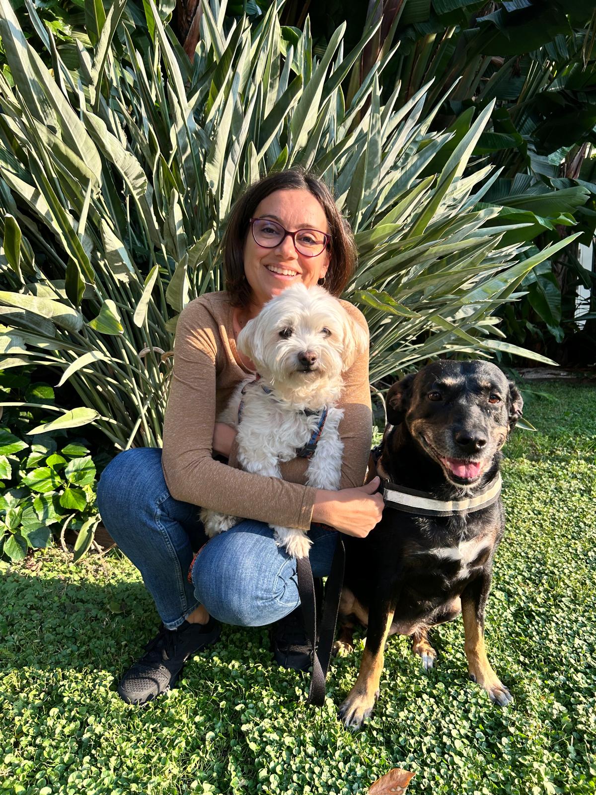 Femme souriante avec lunettes tenant un petit chien blanc, à côté d'un grand chien noir, dans un jardin luxuriant.