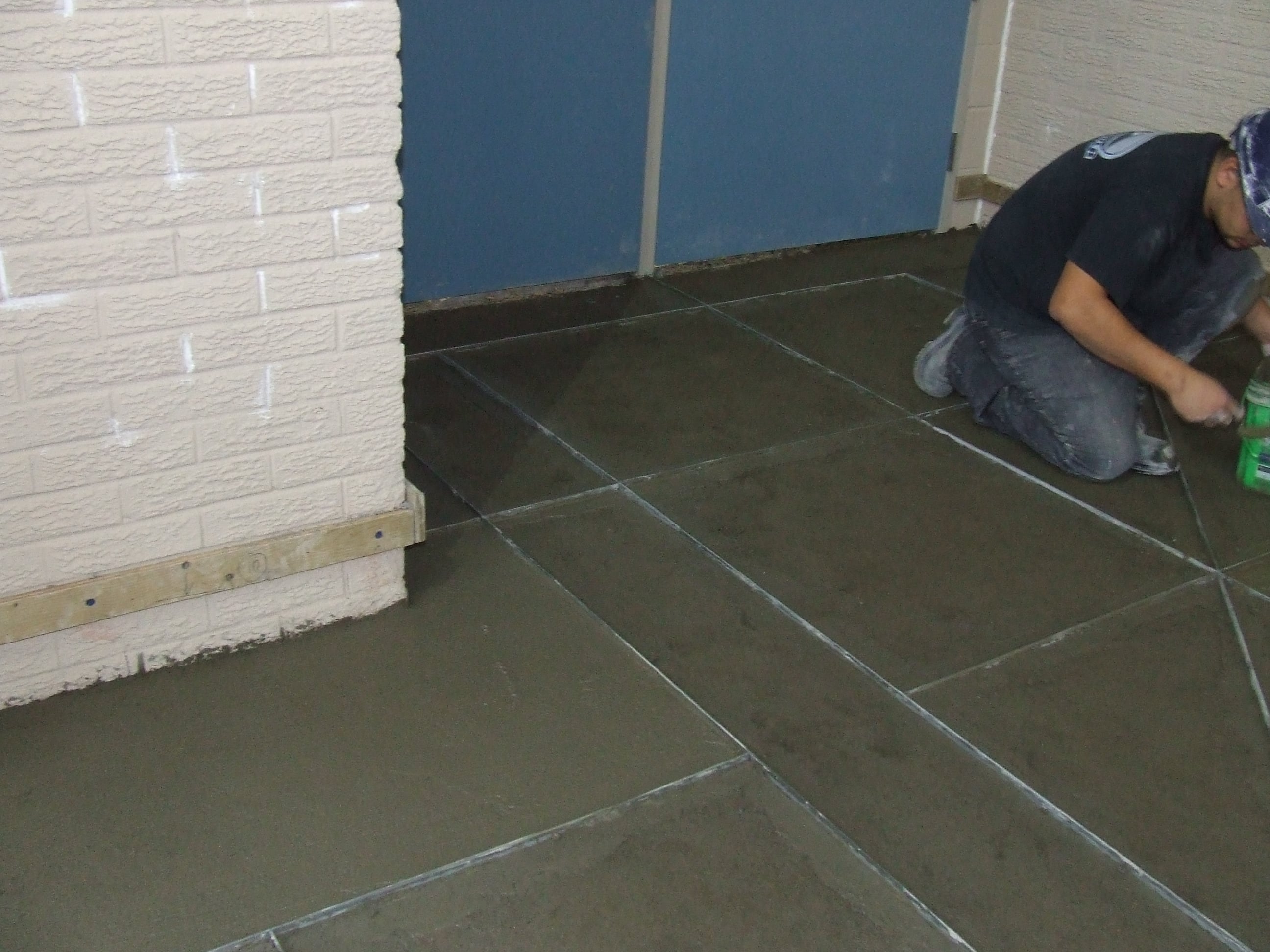 Person in construction attire finishing freshly poured concrete floor near a textured brick wall and blue door.
