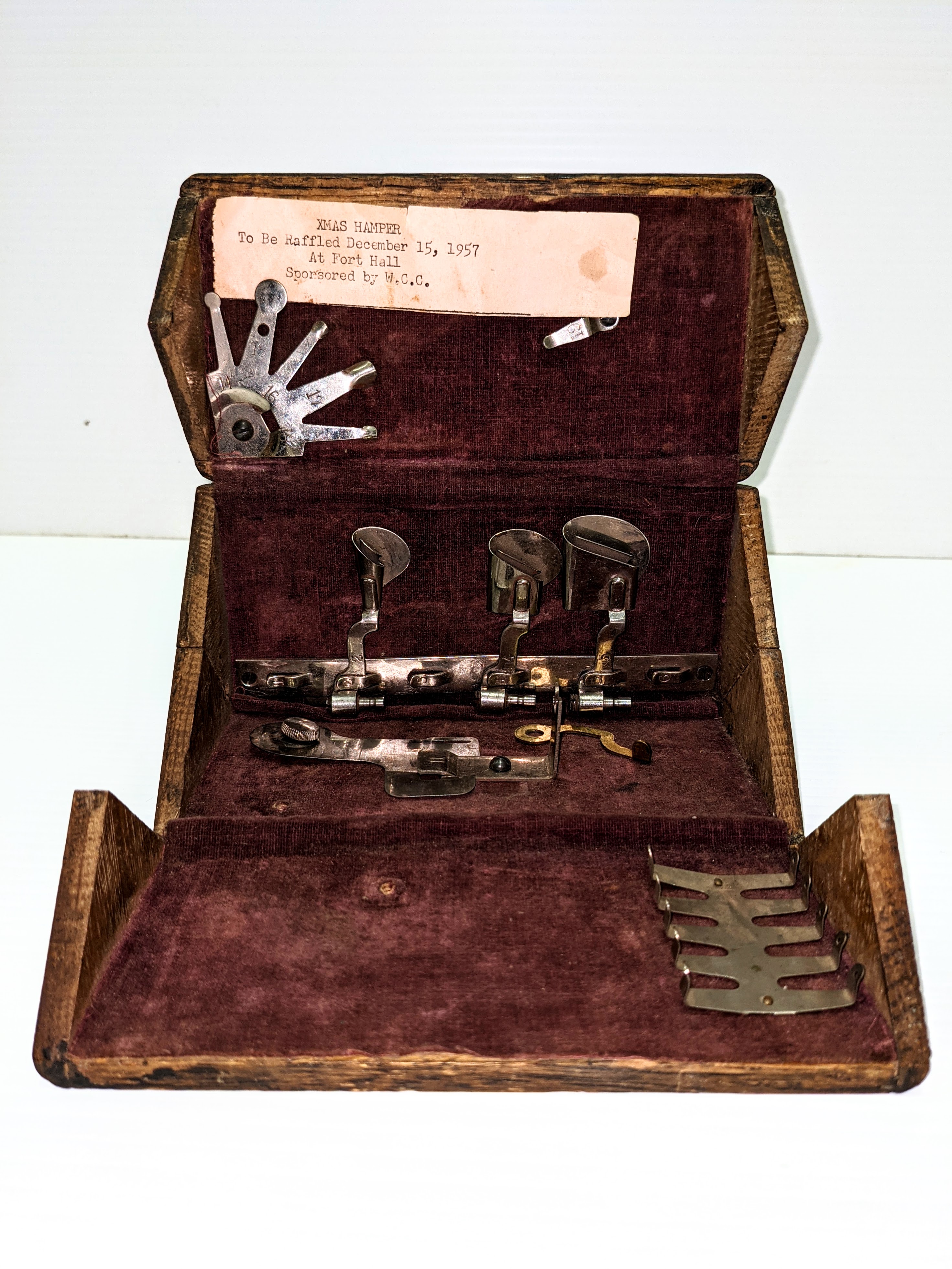 Known as a "Puzzle Box" by modern collectors, this foldable, velvet lined, wooden box is a storage device for various adapters and parts of a Singer sewing machine. First invented in 1889, each box had specific parts for one of 14 models of Singer sewing machine. This box is not complete with it's arrangement of parts and one of the pieces appears to be homemade. Perhaps more interesting is the caption included in the box noting that the unit was donated by the W.C.C (Womens Community Club) to be raffled for a "Xmas Hamper" at the Fort Hall on December 15, 1957.

07/06/2021
2004.11.23.03 / MacRoberts, Estella + Lewis