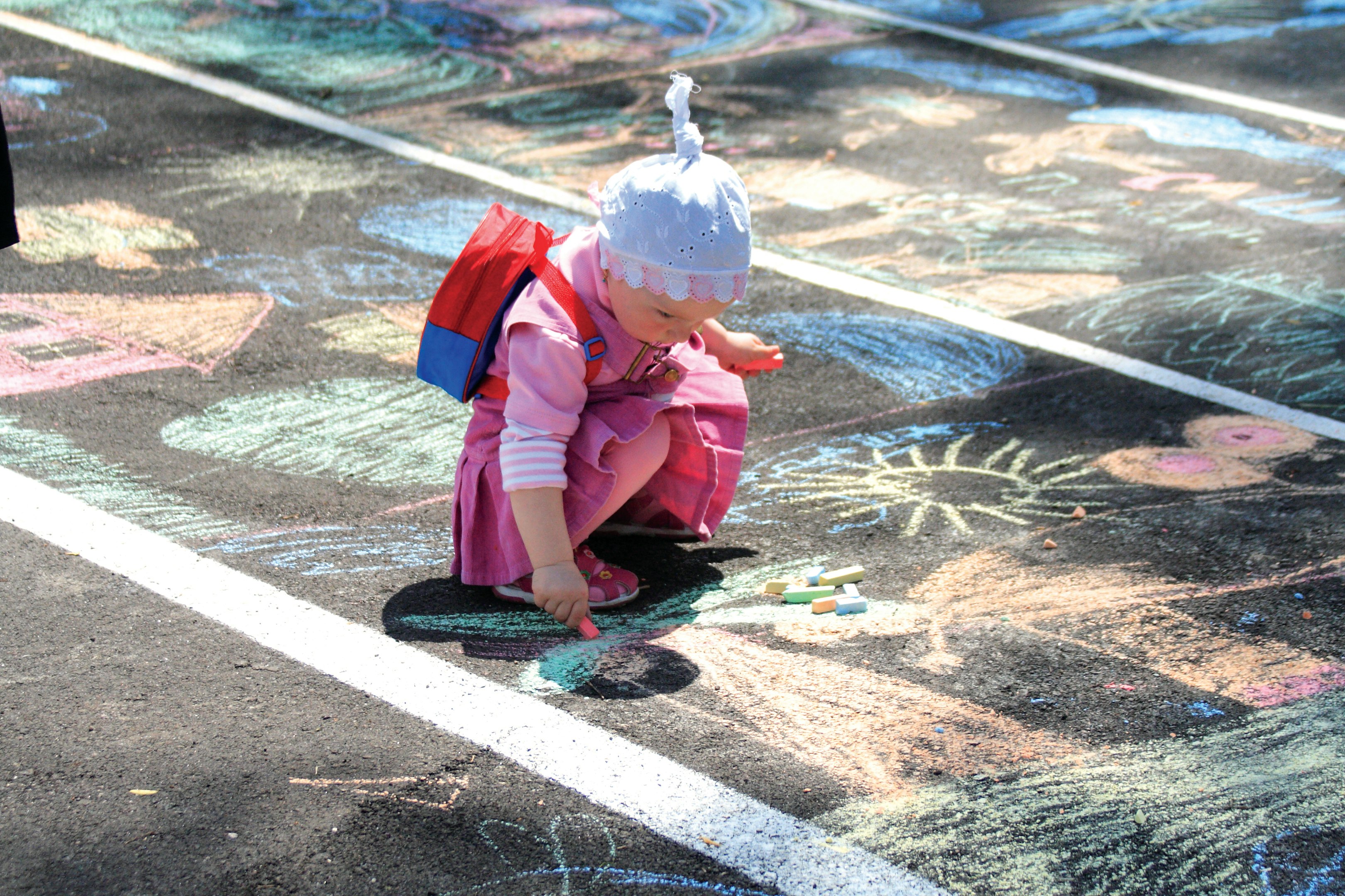 Young child in pink outfit drawing with chalk on asphalt, colorful art surrounding. Young child in pink outfit drawing with chalk on asphalt, colorful art surrounding.