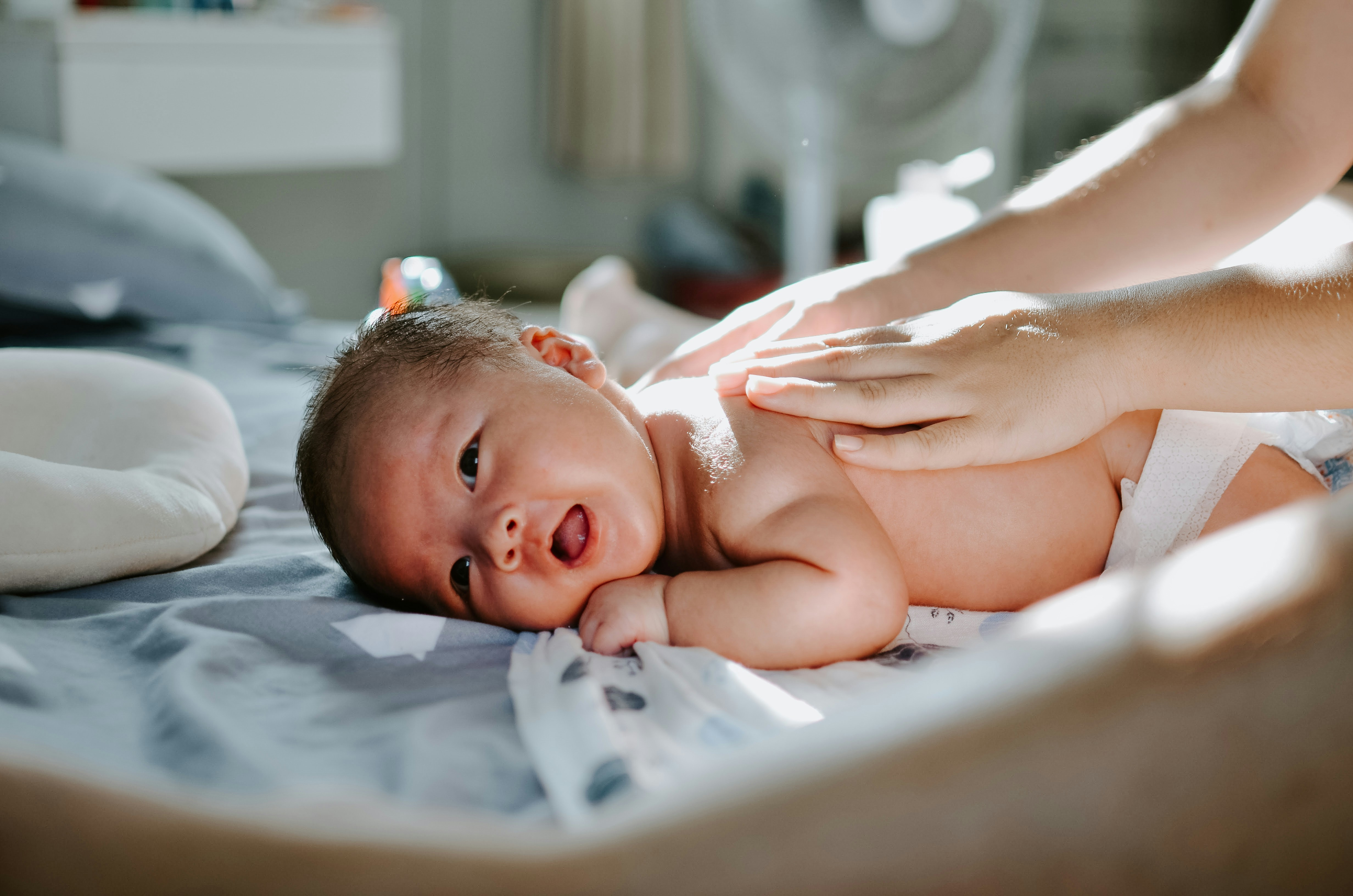 Smiling baby lying on a bed while receiving a gentle massage from an adult hand in a softly lit room.