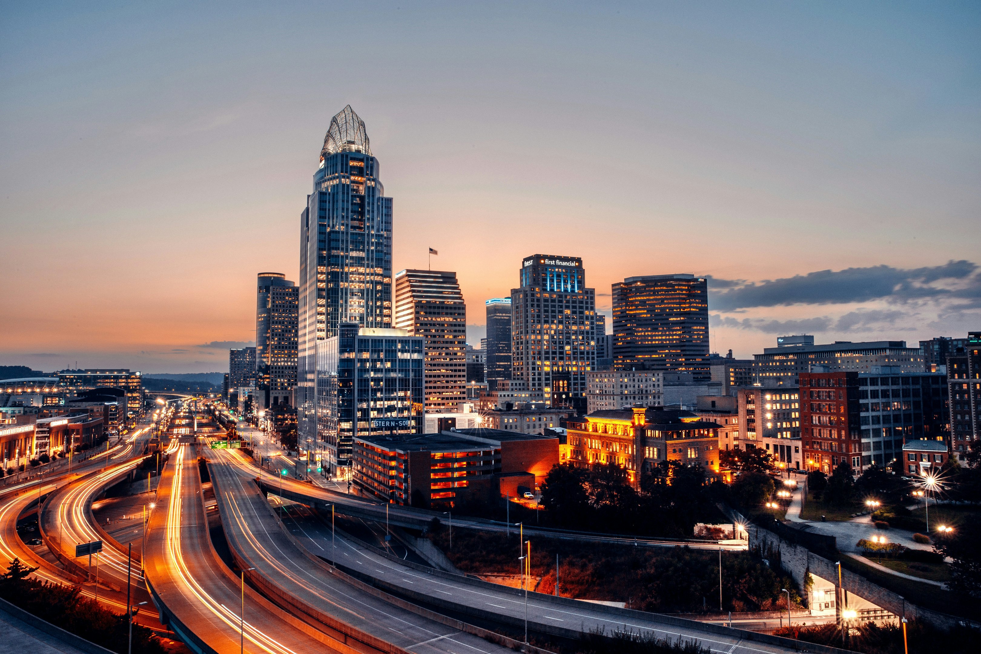 Evening cityscape of downtown Cincinnati skyline with illuminated skyscrapers and highway lights.