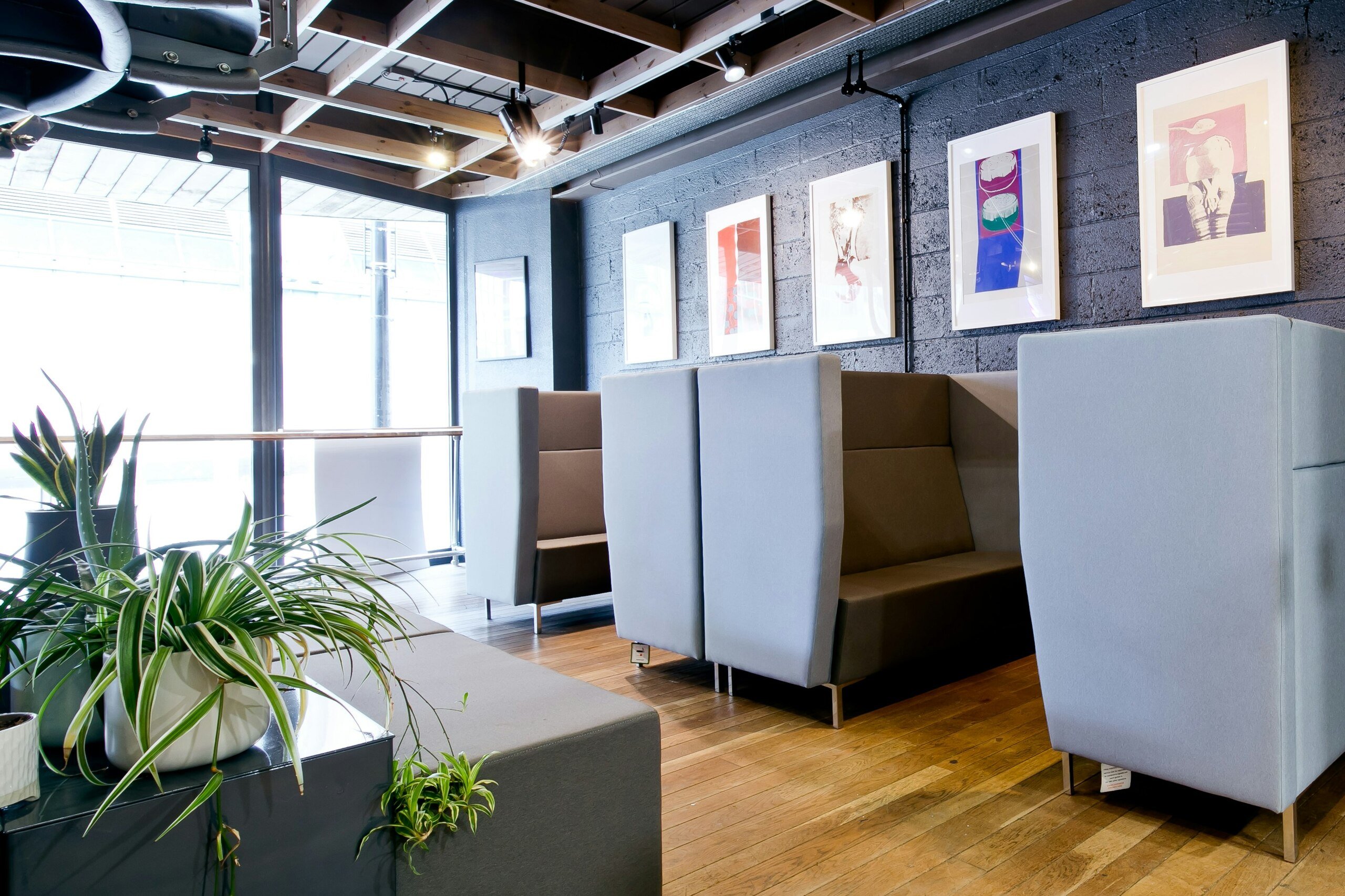 Modern office seating area with grey couches, potted plants, wooden flooring, and framed artwork on a brick wall.
