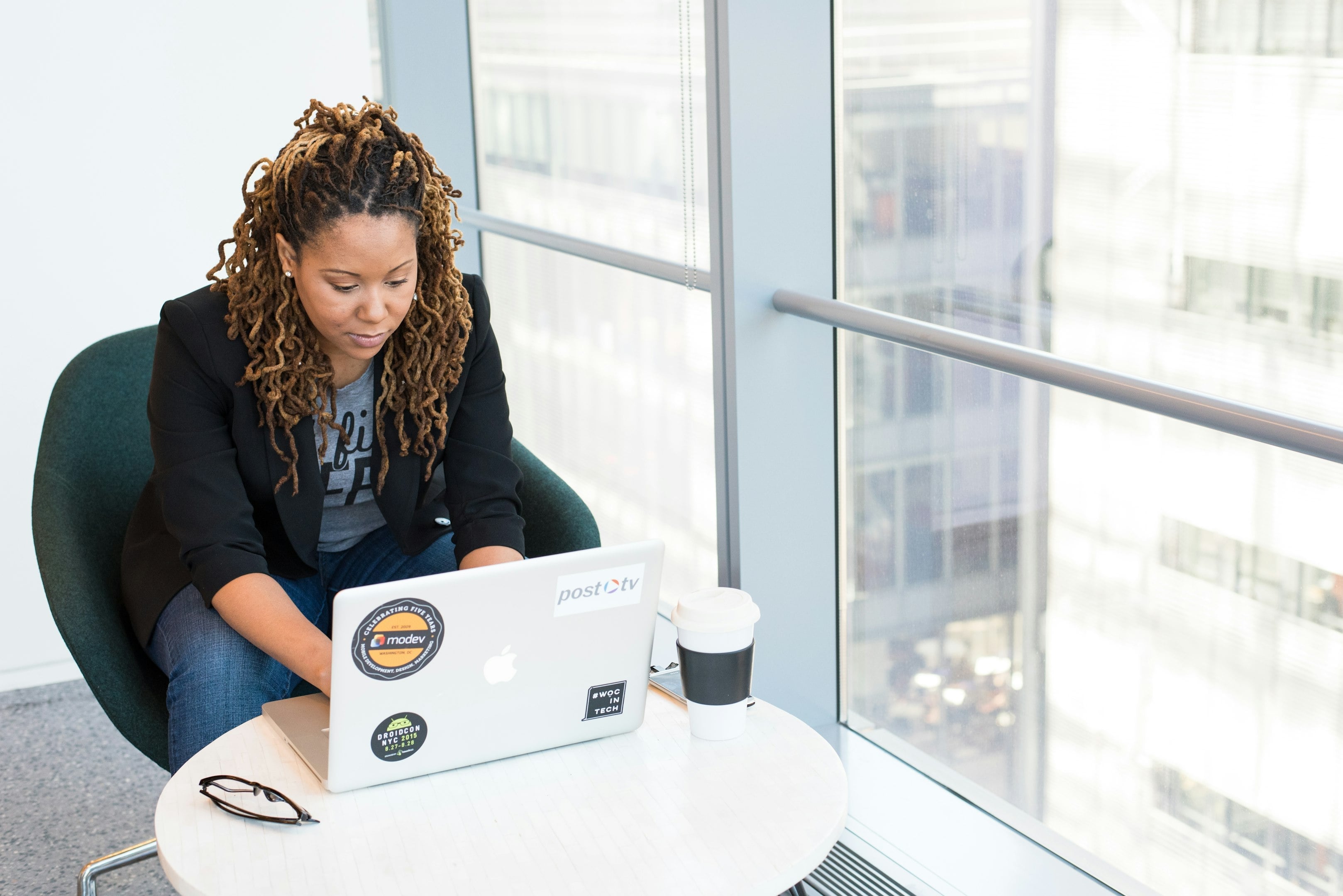 Woman with braided hair working on a laptop at a round table next to a large window in an office setting.
