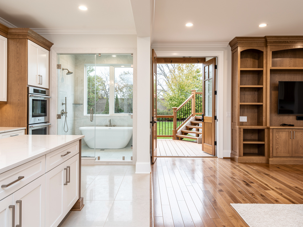 Modern open-concept kitchen and living area with glass shower, freestanding tub, hardwood floors, built-in wood entertainment center, and open doors to deck