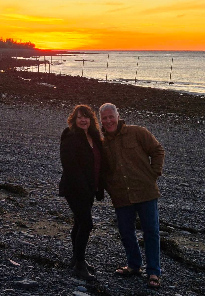 Kerry and her Husband smiling at the camera by the water at sunset.