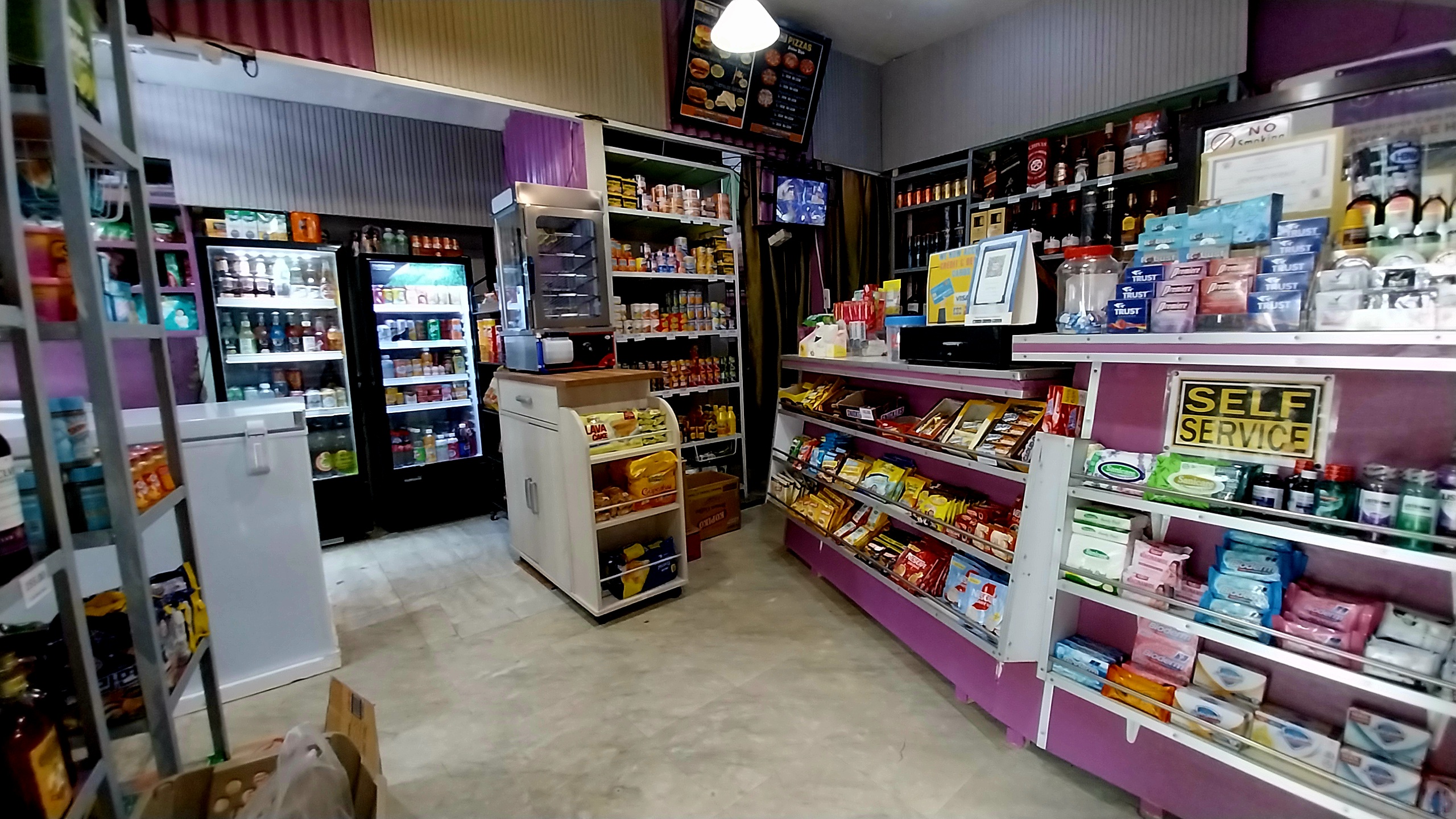 Interior of a small convenience store with shelves stocked with snacks, drinks, and toiletries, featuring a prominent 'Self Service' sign.