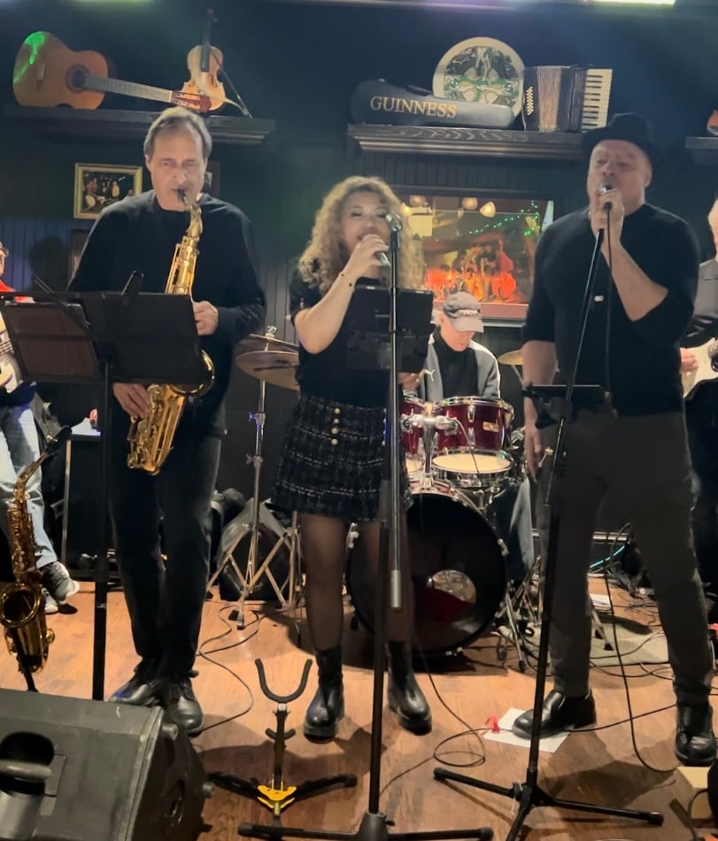 Live band performing on stage with saxophonist, female singer, male vocalist, and drummer in a pub setting