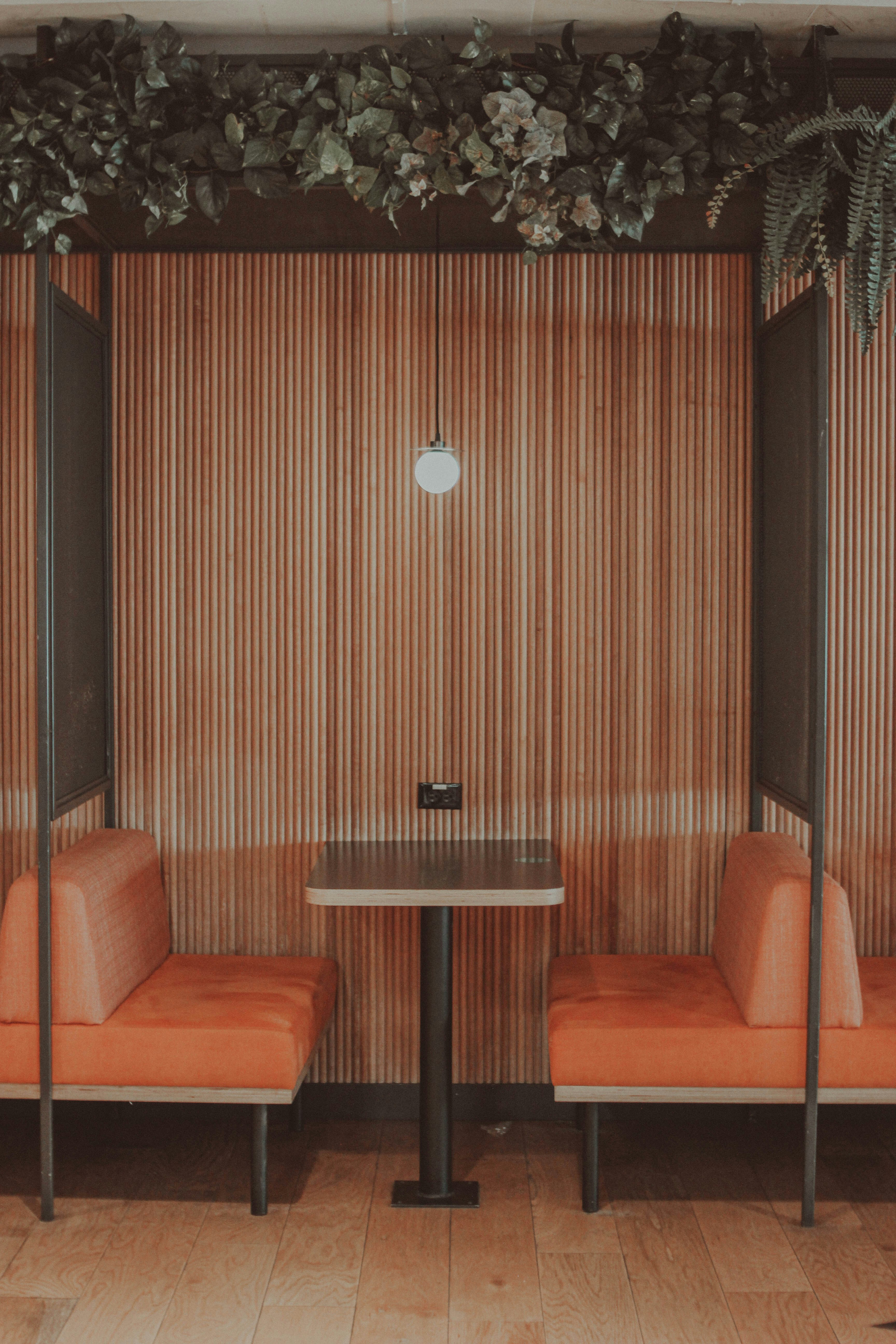 Cabina de restaurante con asientos naranjas, mesa de madera y lámpara colgante, decorado con pared de listones de madera y plantas colgantes.