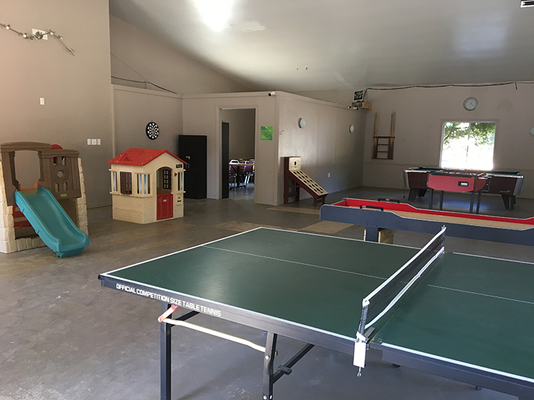 Paysage de camping en montagne avec tentes, rivière et canoë, entouré de forêts et montagnes sous un ciel bleu.