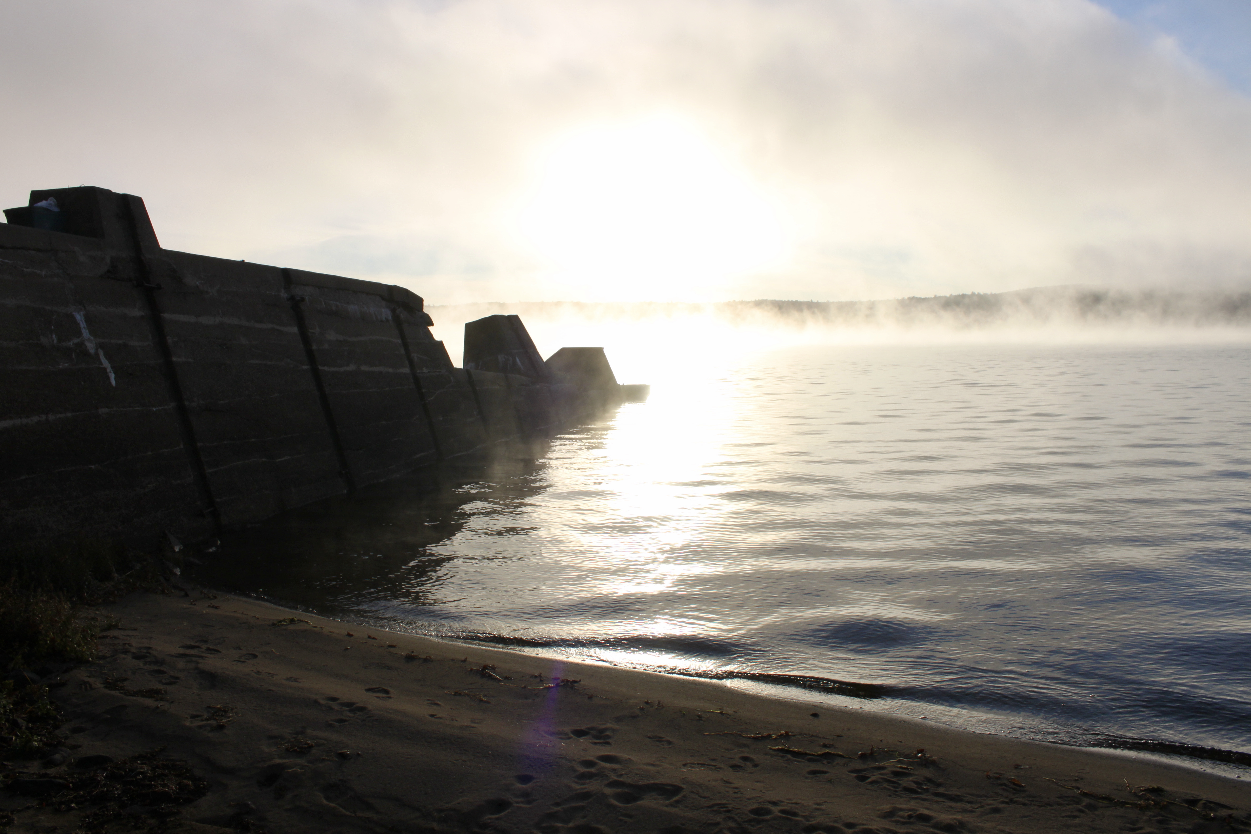 Scenic view of a concrete breakwater at a misty lake shore during sunrise, with gentle waves lapping against the sand.