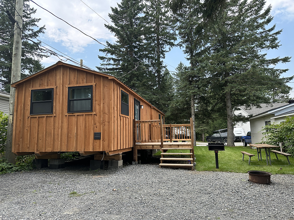 Camping en montagne avec tentes, cabane en bois, rivière, arbres verts et montagnes en arrière-plan par une journée ensoleillée.