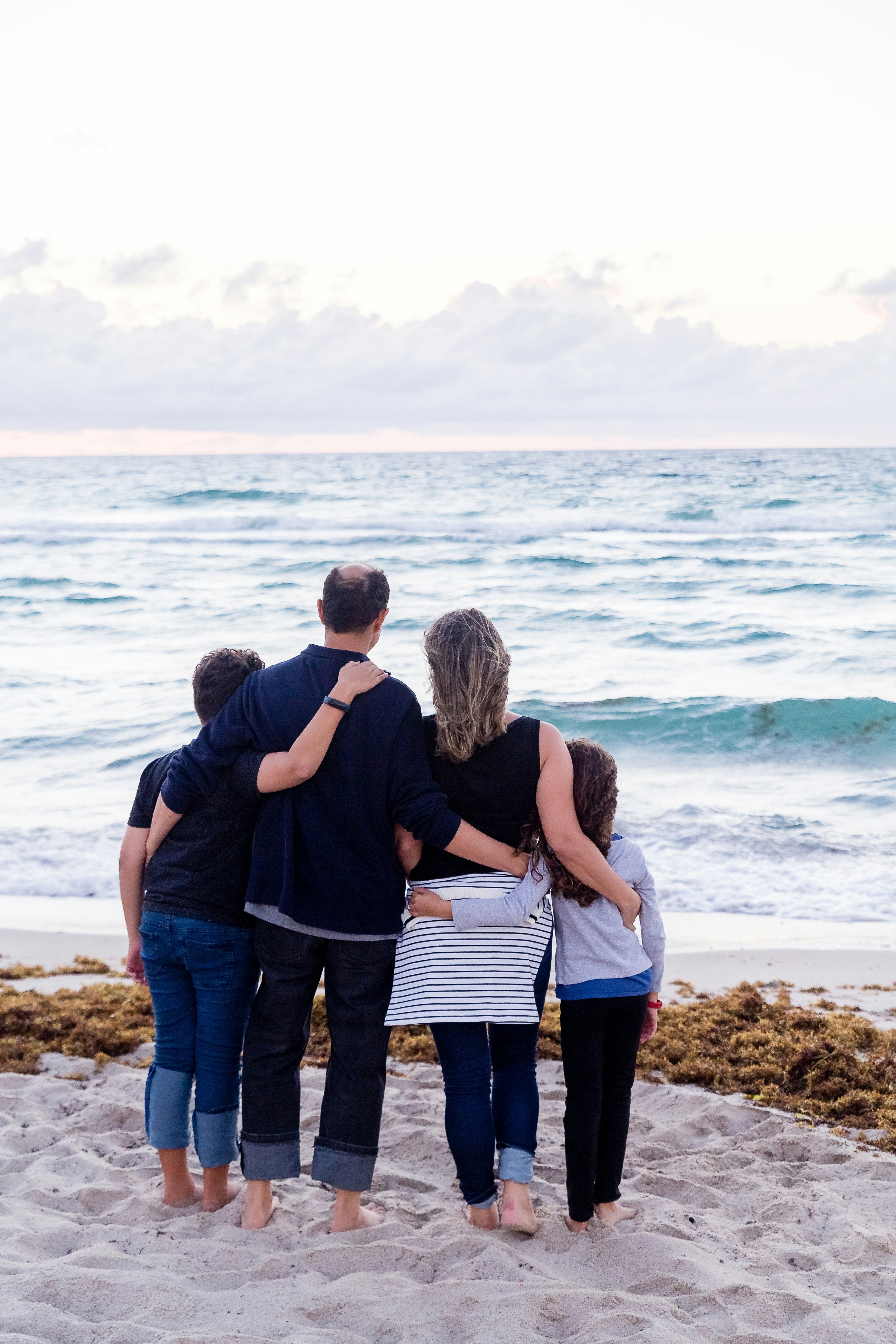Family standing arm in arm on sandy beach watching ocean waves at sunset
Family standing arm in arm on sandy beach watching ocean waves at sunset