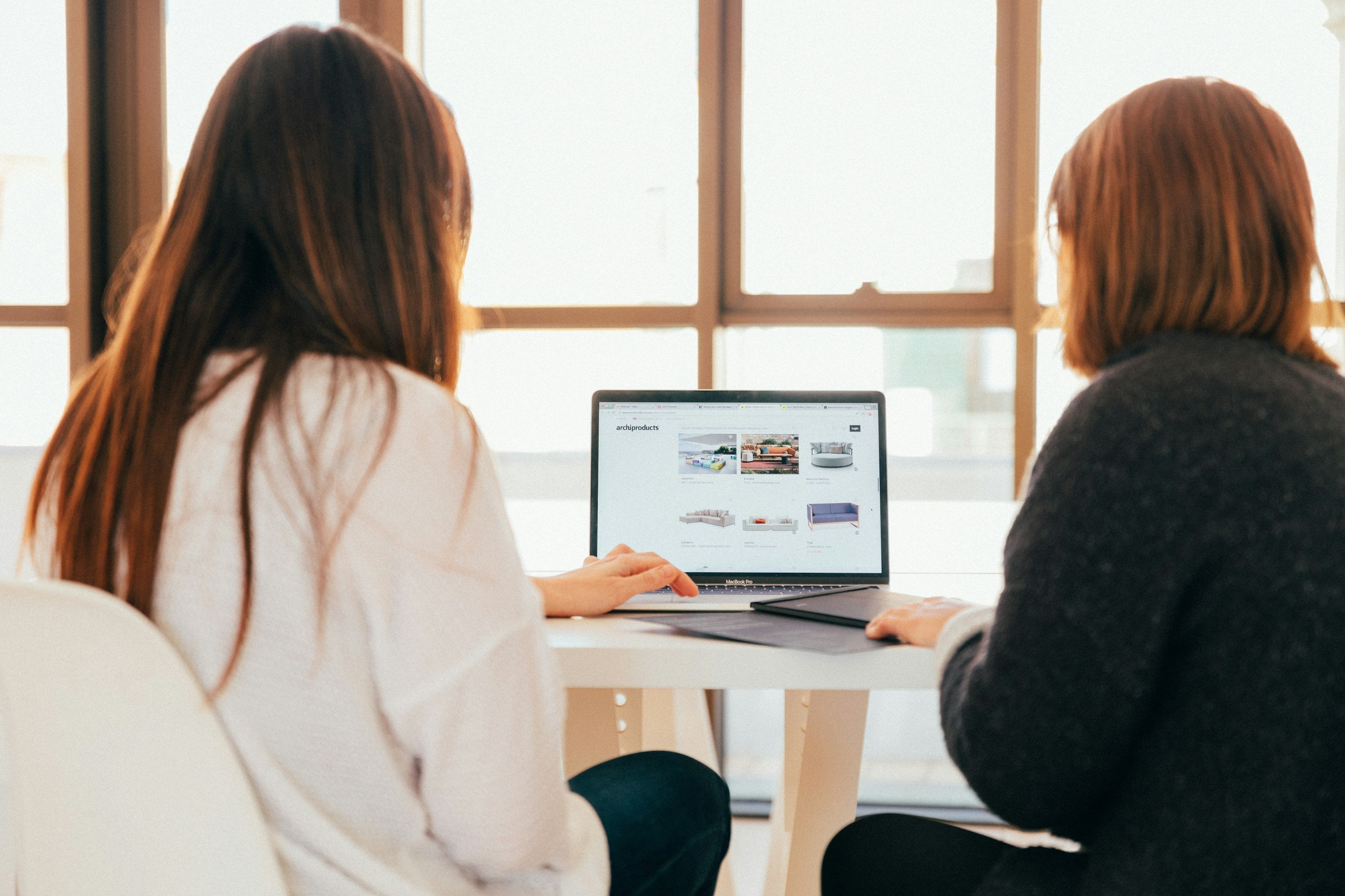 Two women sitting at a table using a laptop displaying a website with furniture images, in a bright room with large windows.