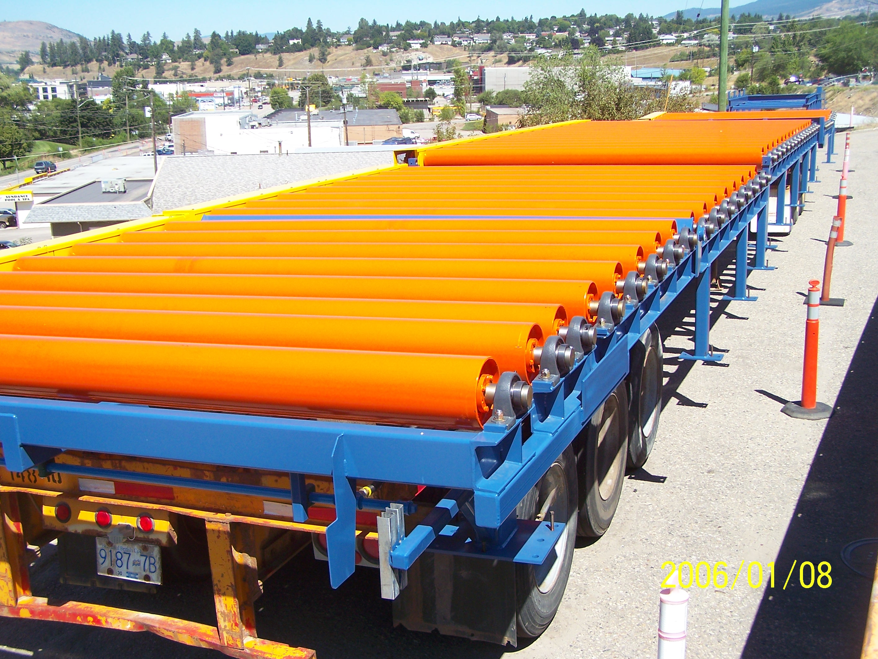 Long flatbed truck loaded with bright orange cylindrical pipes on a blue frame, parked in an industrial area with buildings and hills in the background.
