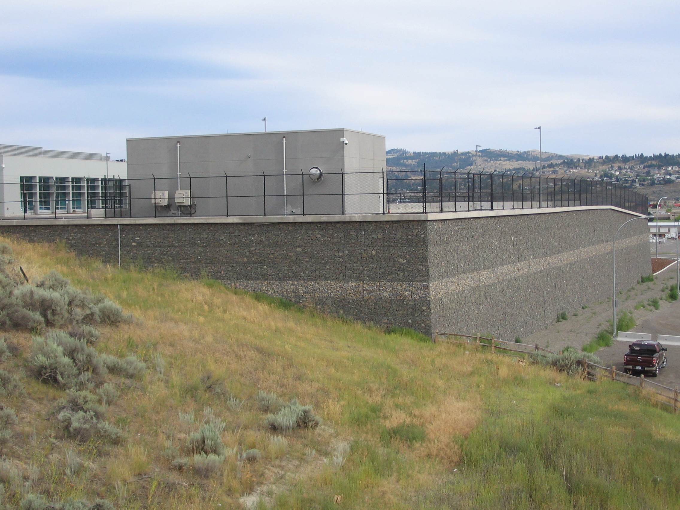 Large stone retaining wall with a fenced facility on top, surrounded by grassy landscape and distant hills.