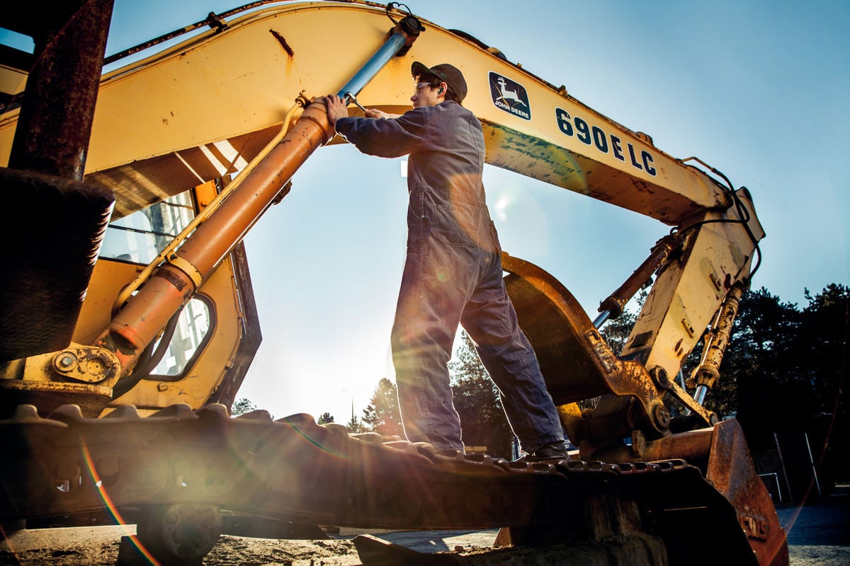 Construction worker repairing hydraulic arm on yellow John Deere excavator at outdoor worksite