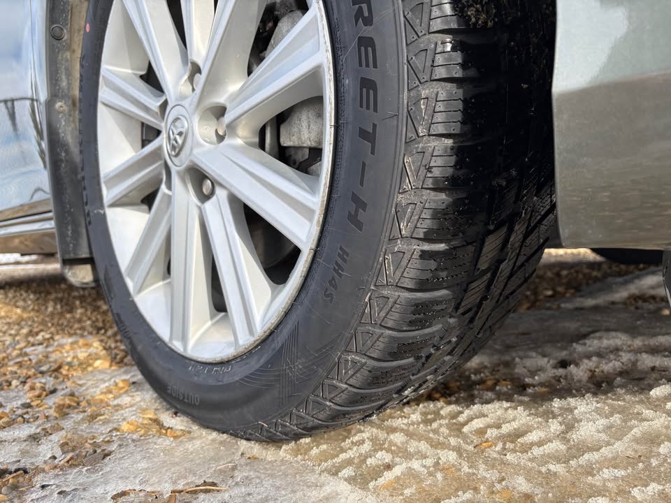 Close-up of a car wheel with winter tire tread on a snowy and gravel surface.