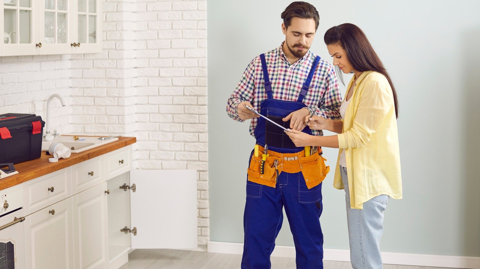 Female homeowner consulting handyman in blue overalls with tablet in modern kitchen setting.