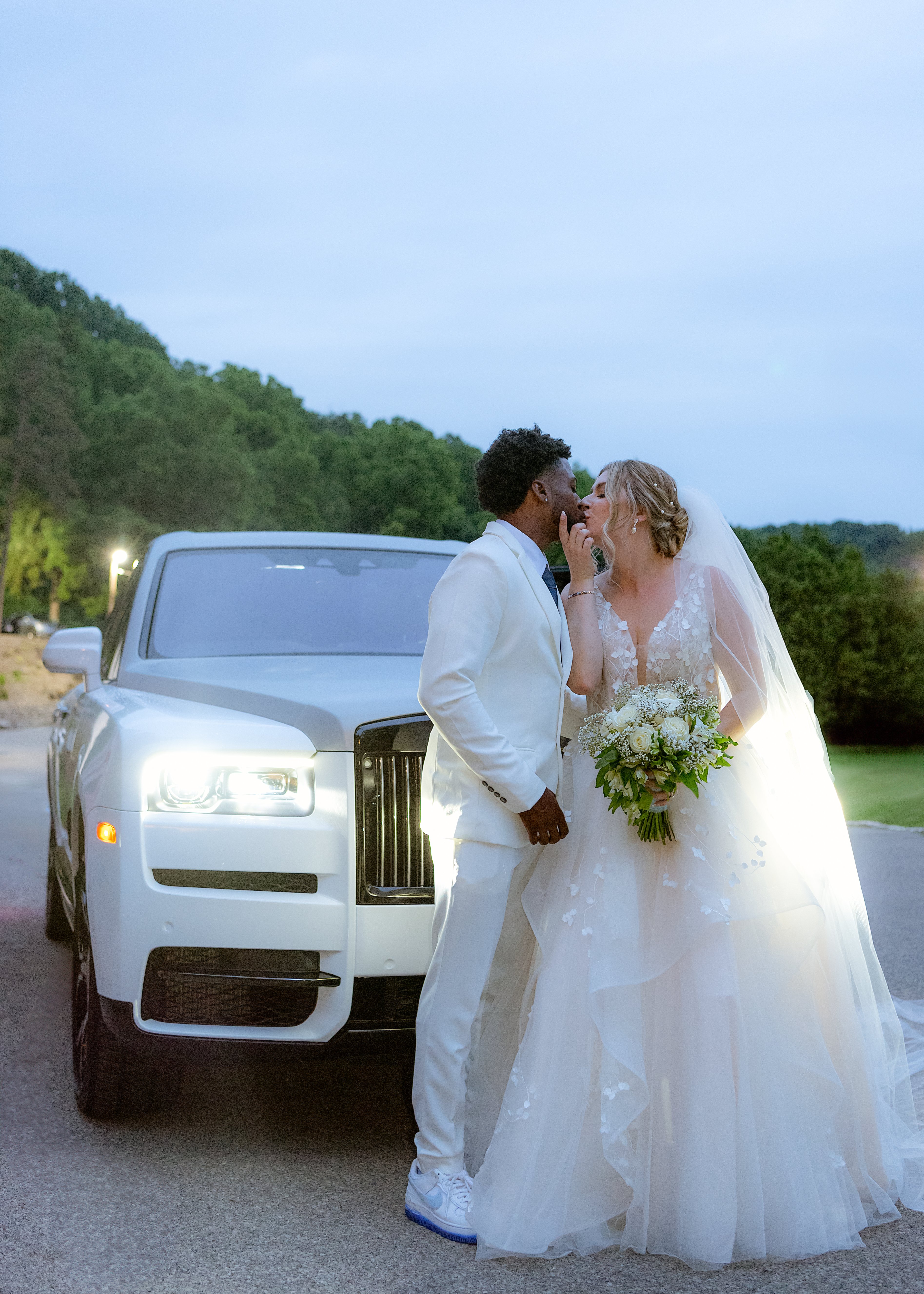 Bride and groom in elegant attire kissing beside a luxury white car with green scenic background.