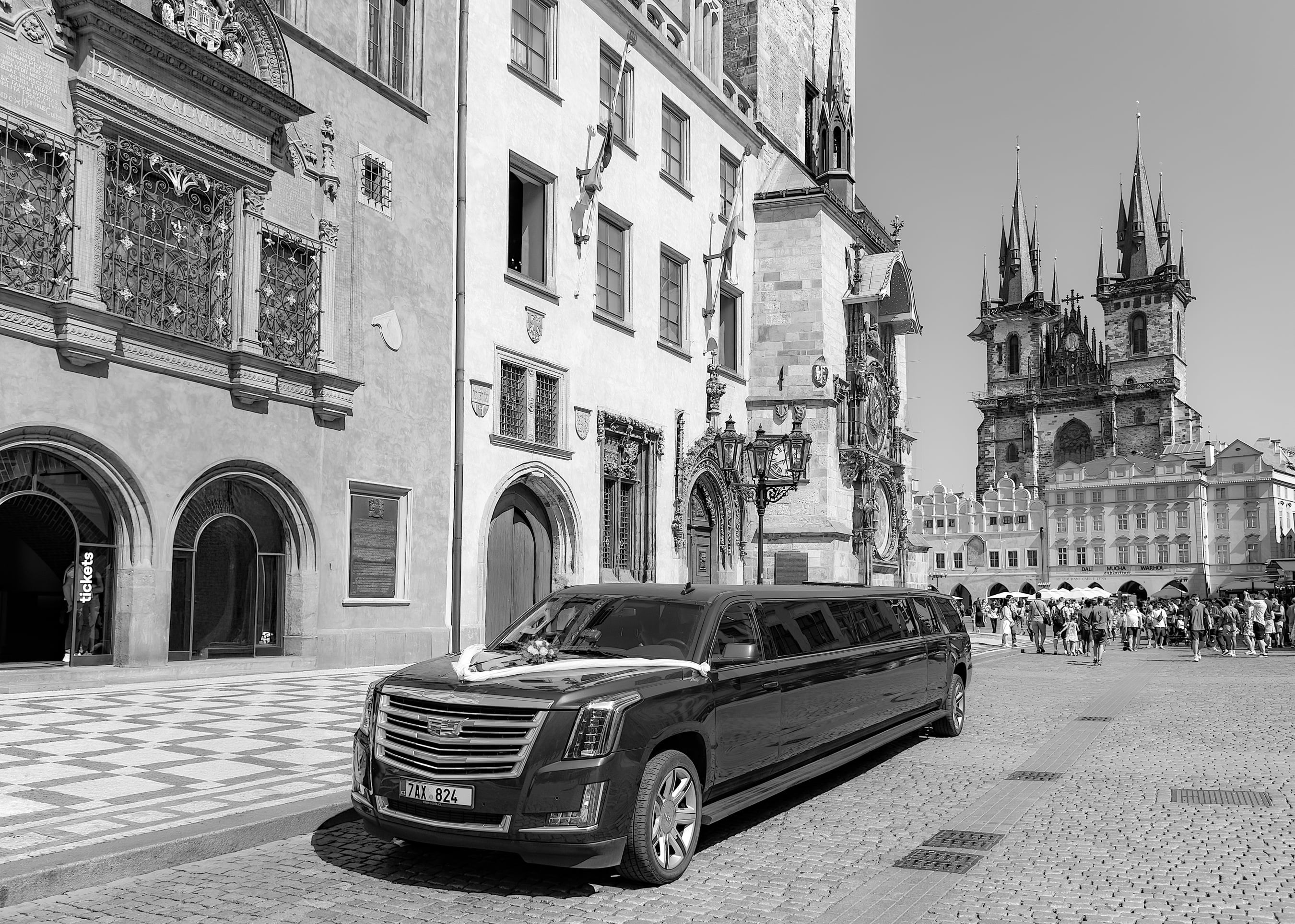 Black limousine parked on cobblestone street in historical European city with Gothic cathedral and ornate architecture in background, black and white photo.