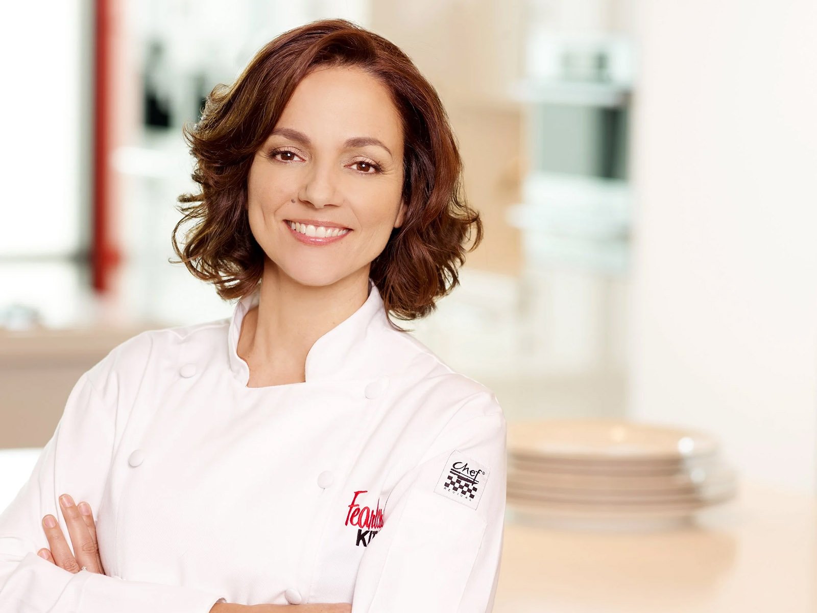Smiling female chef in a modern kitchen with wooden shelves and plants.