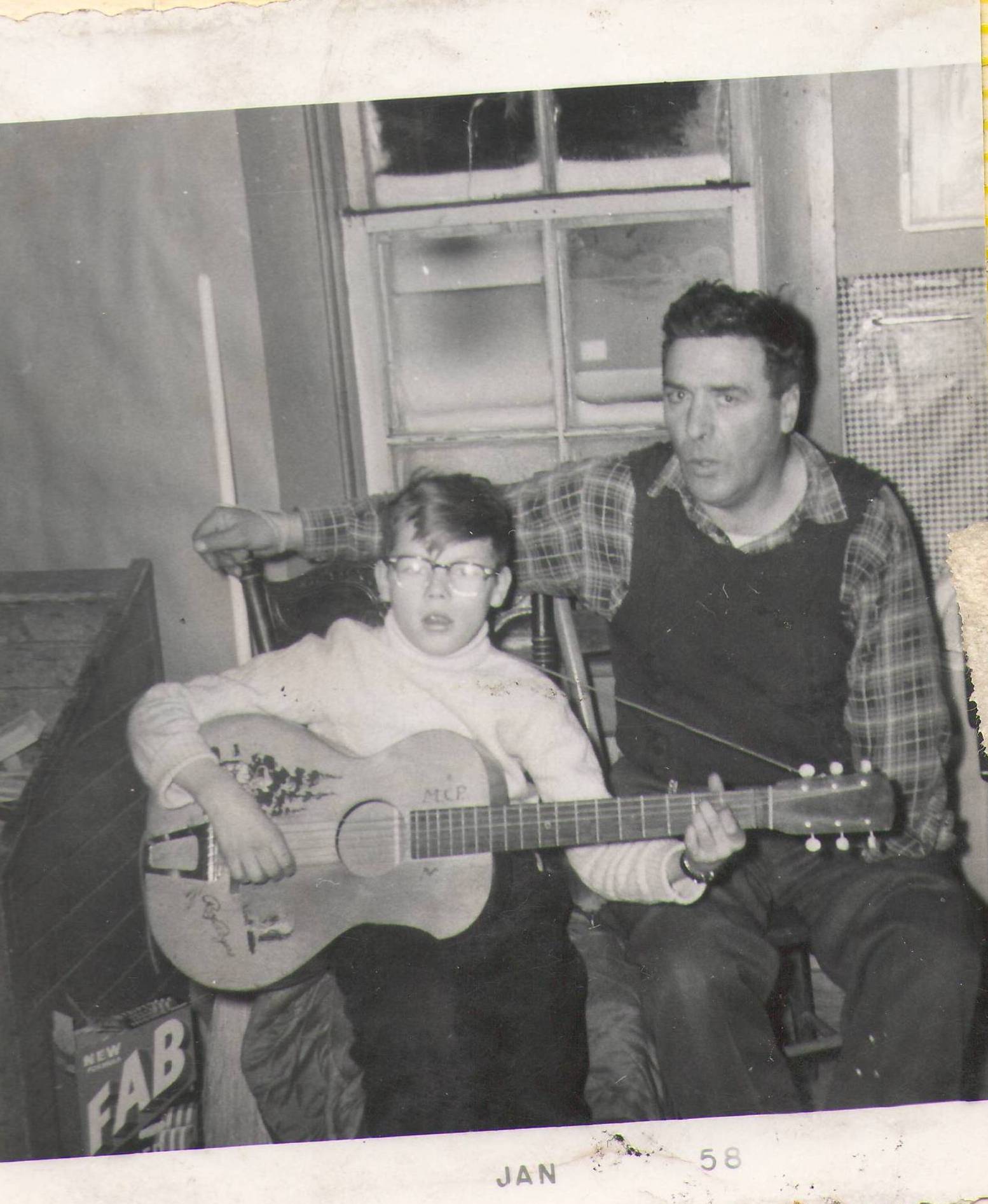 This picture comes from the Maria Lizotte collection and the young lad playing the guitar is labelled as Clifford Pettman - we are hoping you can help identify the older gentleman! Note the date at the bottom of the photo is "Jan 58"

2019.24.628 
 Lizotte Maria