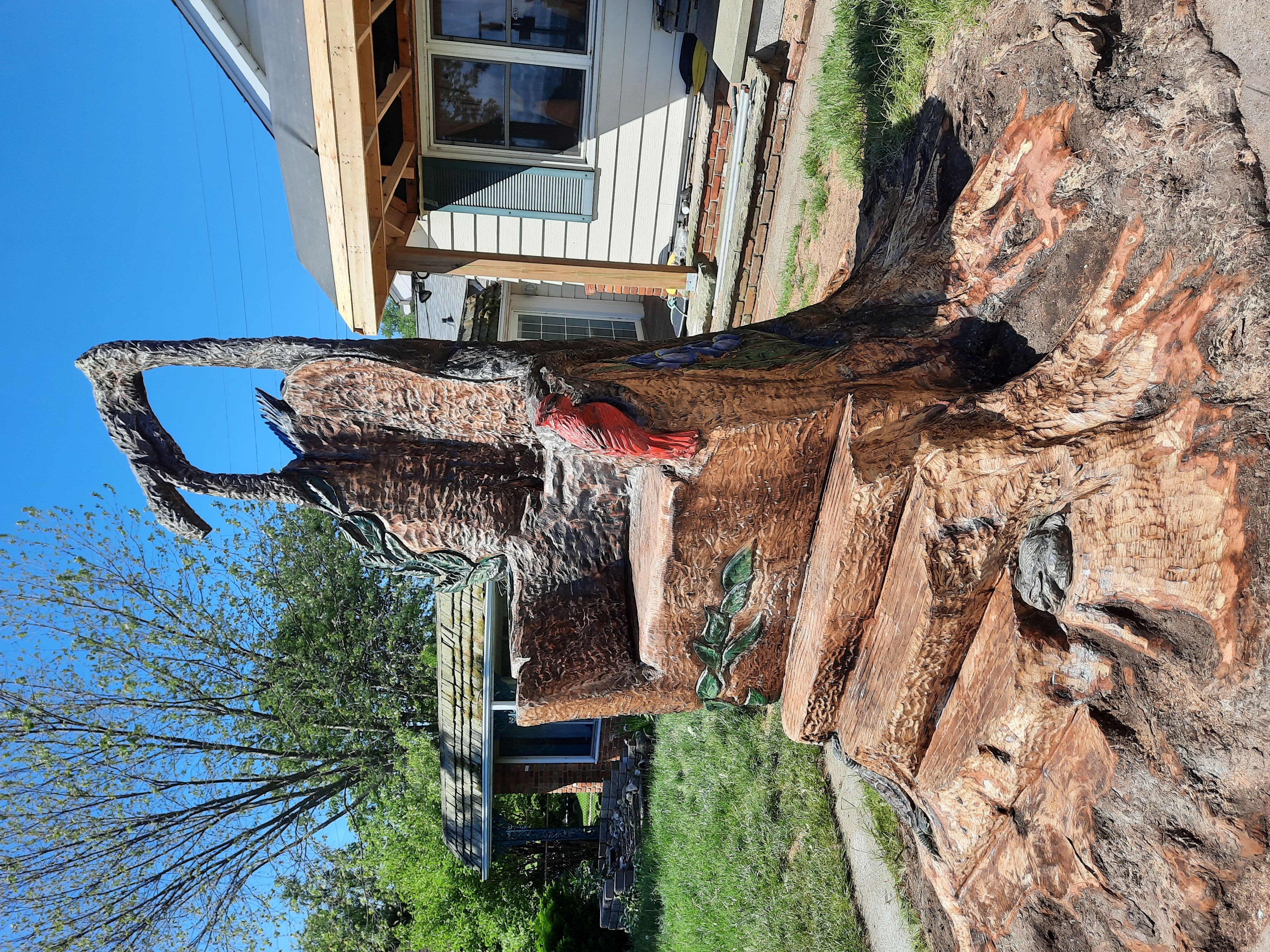Two wood carvers using chainsaws to sculpt a large tree stump into wooden animal statues in a backyard garden