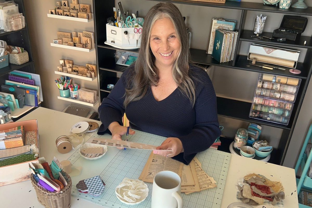 The maker behind Created Me Creative smiling at her craft table, surrounded by handmade journals, paper crafting supplies, and creative projects.