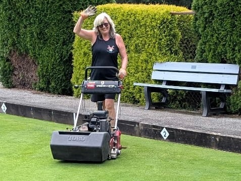 Woman mowing lawn on sports field with hedge and bench in background. Woman mowing lawn on sports field with hedge and bench in background.