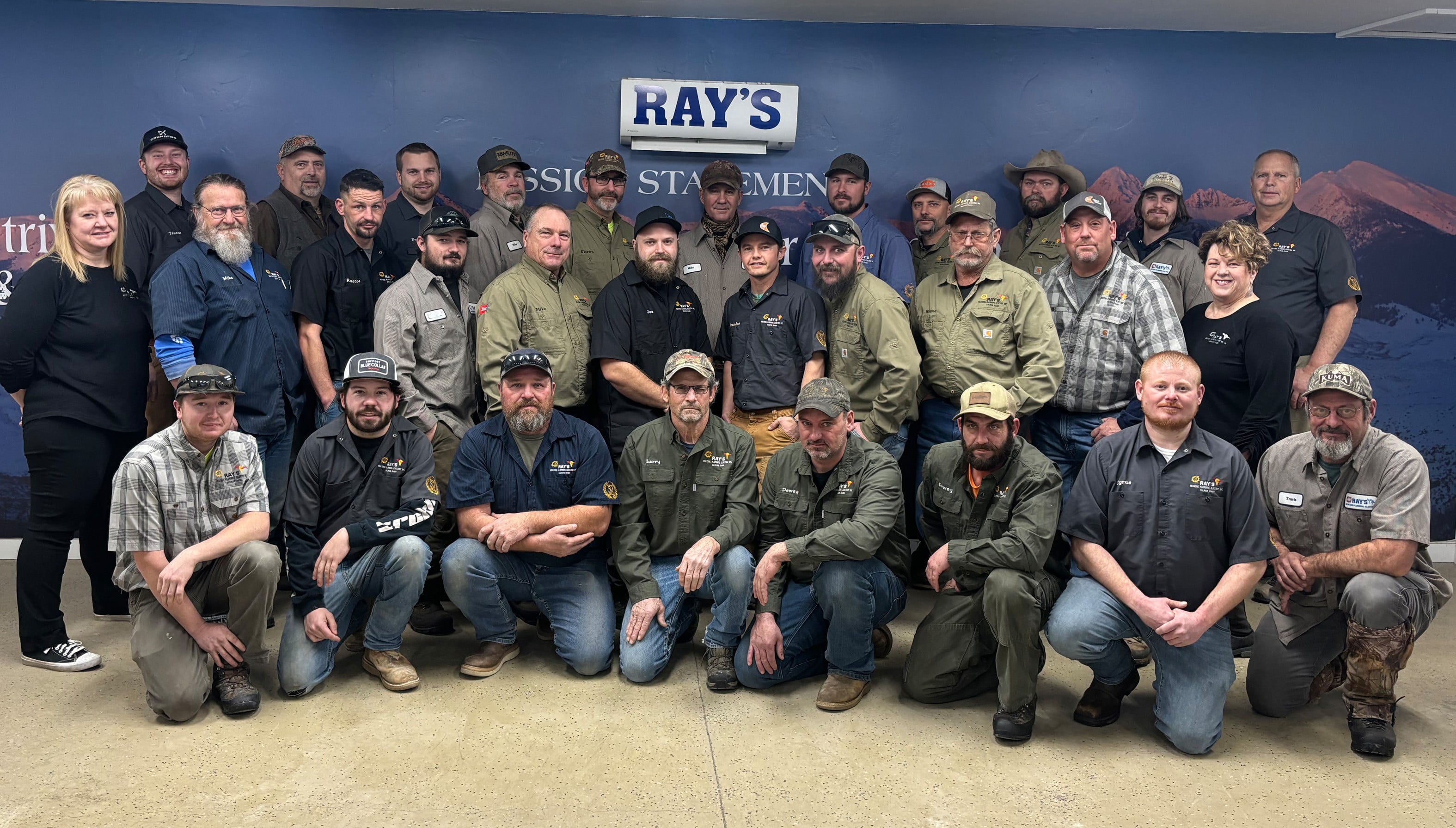 Group of employees in casual workwear posing in front of a blue wall with Ray's logo.