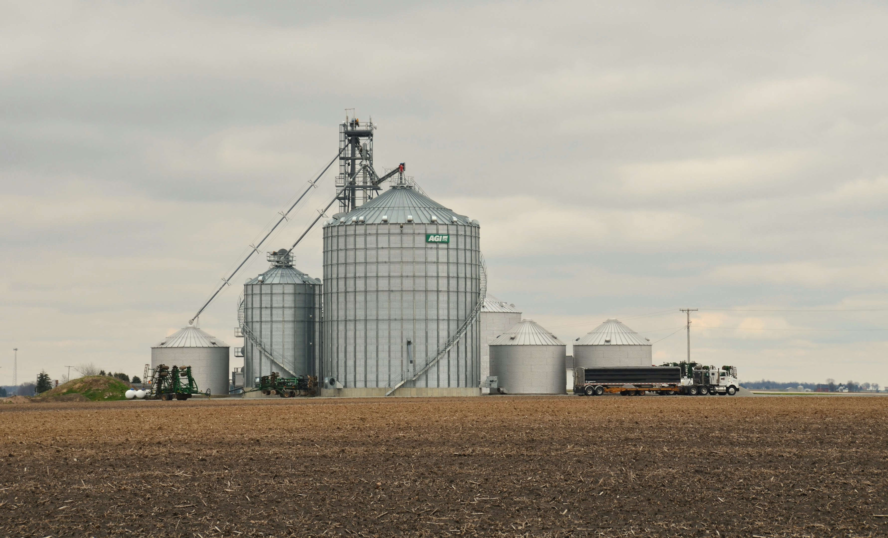 Large agricultural silos and a truck in a rural farmland setting under a cloudy sky.