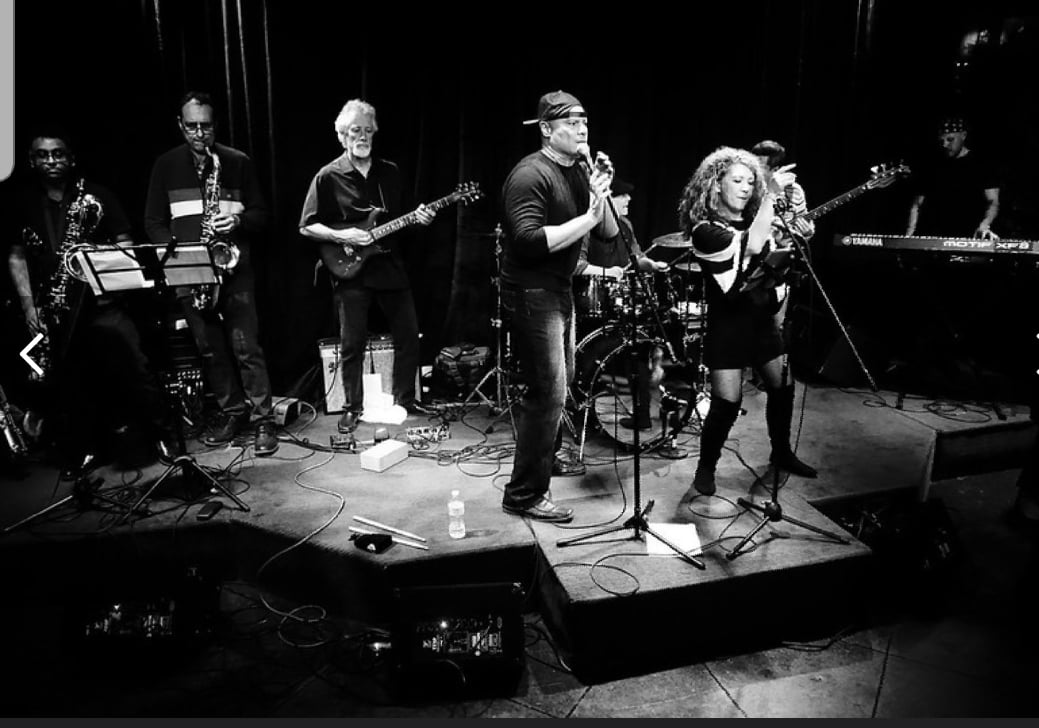 Black and white photo of a live band performing on stage with singers, guitar, saxophones, drums, and keyboard