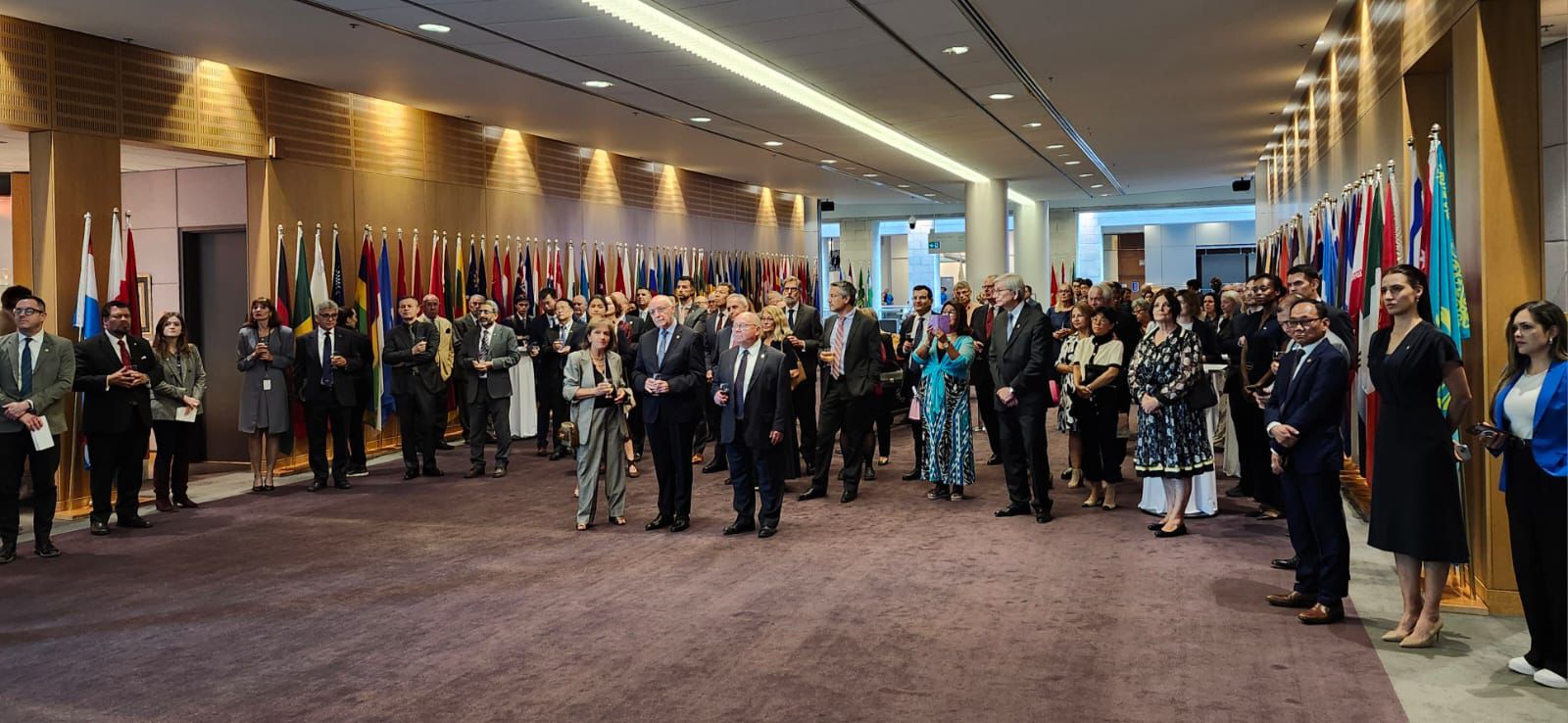 A diverse group of people formally dressed, gathered in a hallway decorated with numerous international flags.
