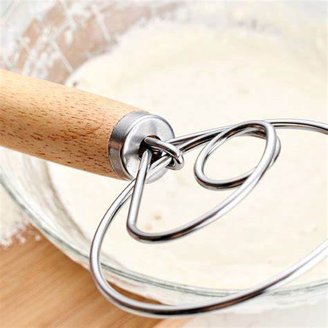 Close-up of a dough whisk with a wooden handle over a mixing bowl containing flour and dough.