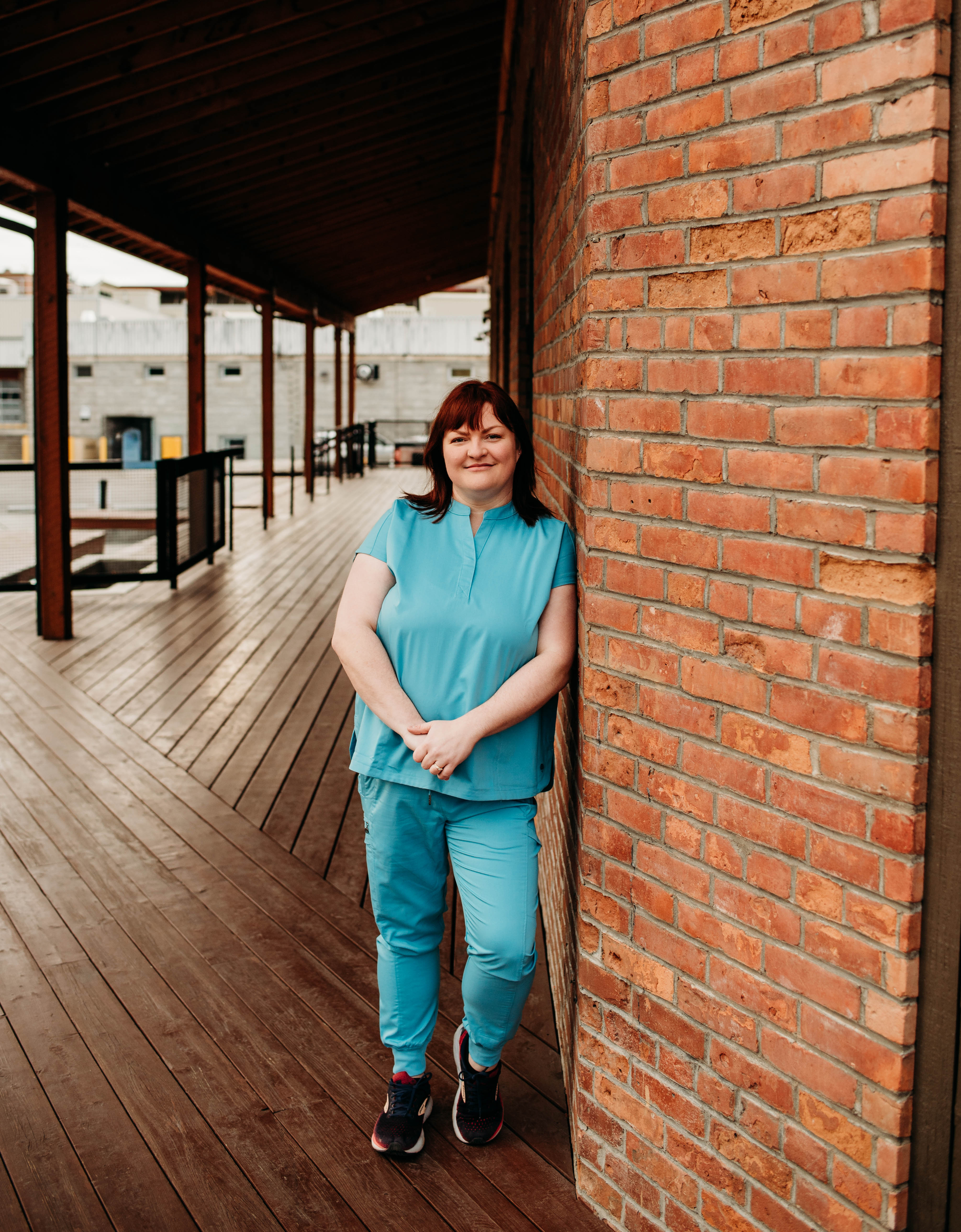Person in blue medical scrubs leaning against a brick wall with a wooden walkway and industrial background.