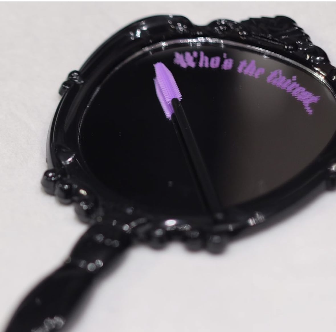 Decorative black handheld mirror with engraved text 'Who's the Queen?' and a purple mascara wand on the reflective surface.