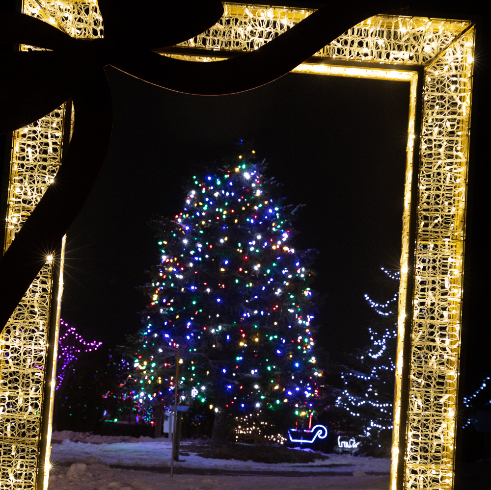 Snow-covered street with Christmas lights, a lit tree, and decorated cafes at night.
