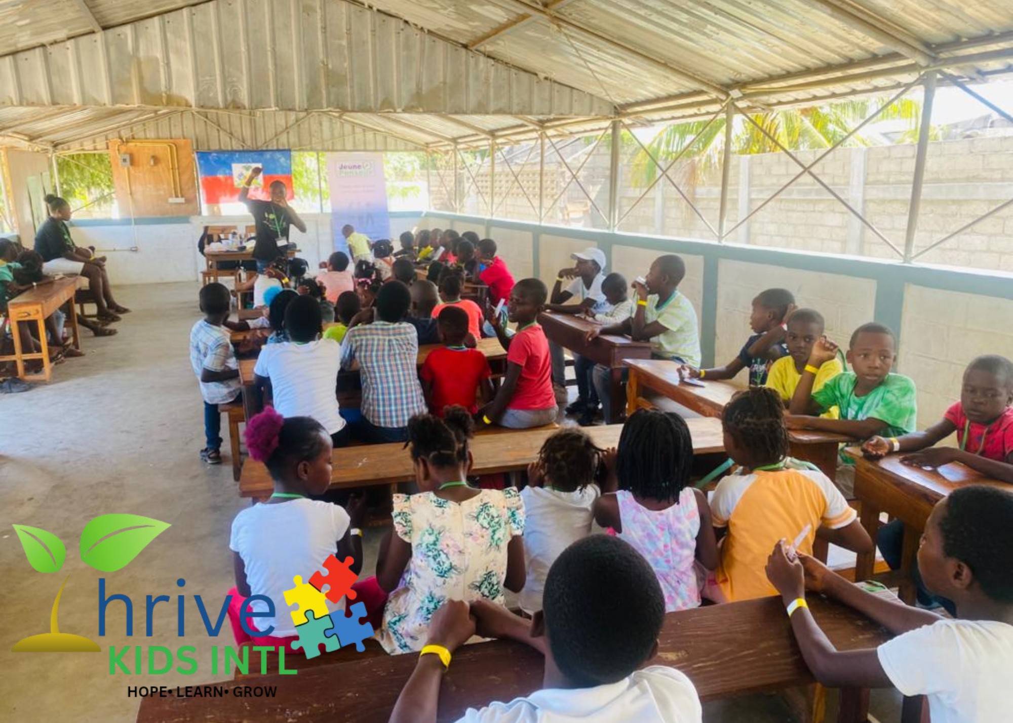 Young children engaged in a classroom activity, drawing and writing at a wooden table with colorful supplies and plants in the background.