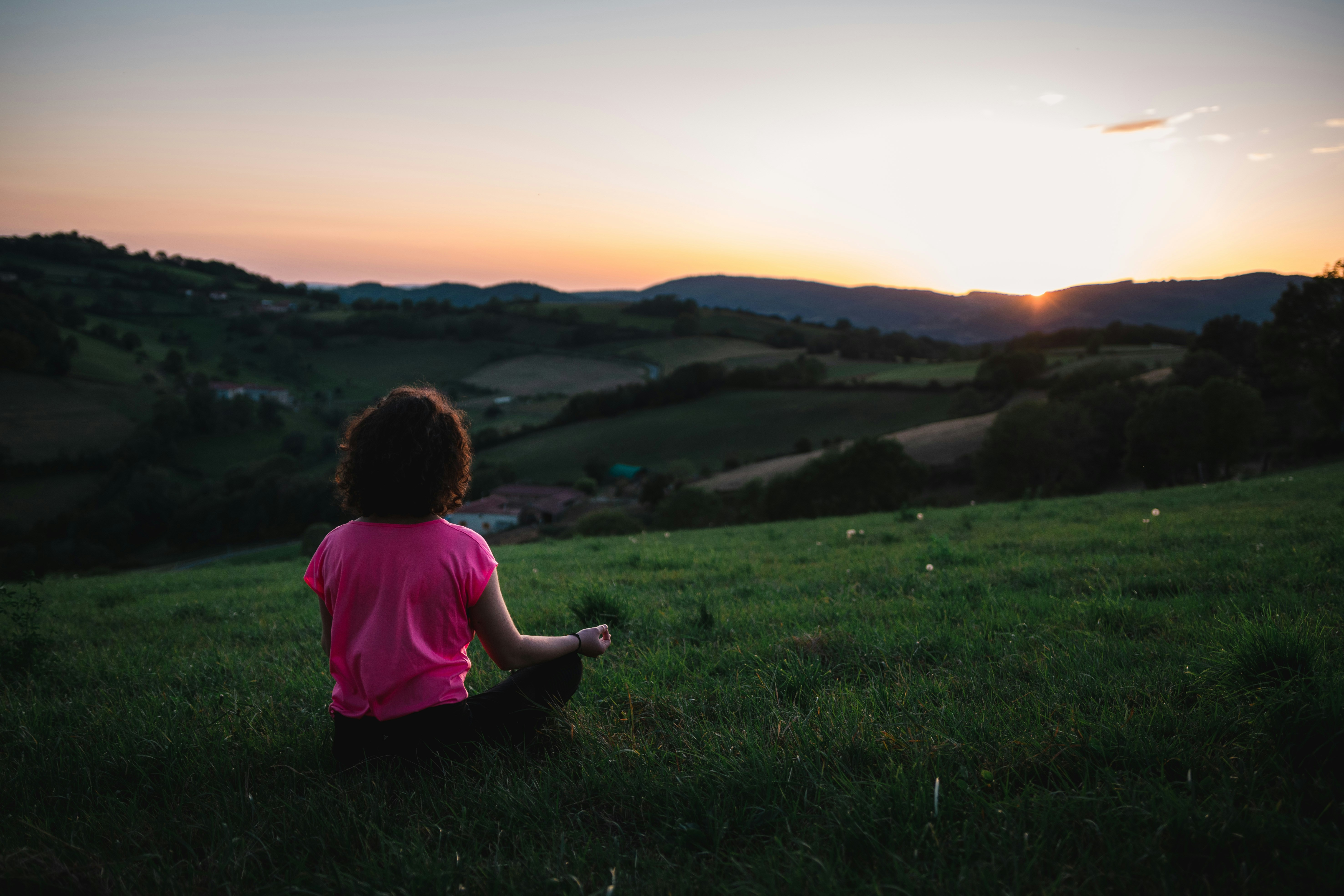Person in pink shirt meditating on grassy hill during sunset overlooking serene landscape with distant hills.