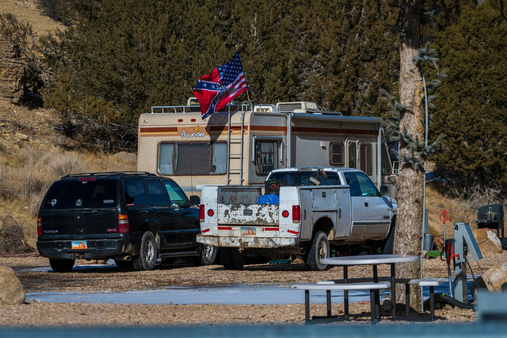 Two parked vehicles, including a black SUV and a white pickup truck, next to a beige RV with flags on it, set in a forested campsite.
