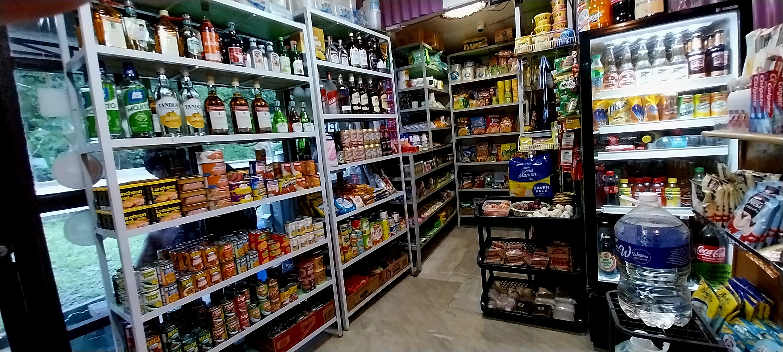 Interior of a small grocery store with shelves stocked with various canned goods, beverages, snacks, and household items.