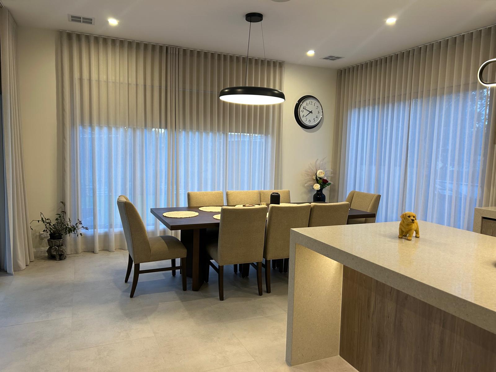 Modern dining room with beige chairs around a wooden table, large windows with sheer curtains, and a kitchen island with a small dog sculpture. Modern dining room with beige chairs around a wooden table, large windows with sheer curtains, and a kitchen island with a small dog sculpture.