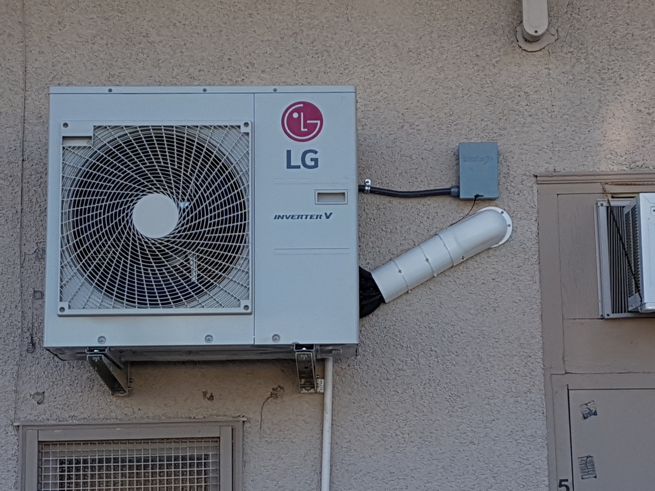 Outdoor air conditioning unit on patio with brick wall and large window, surrounded by plants and cozy blanket.
