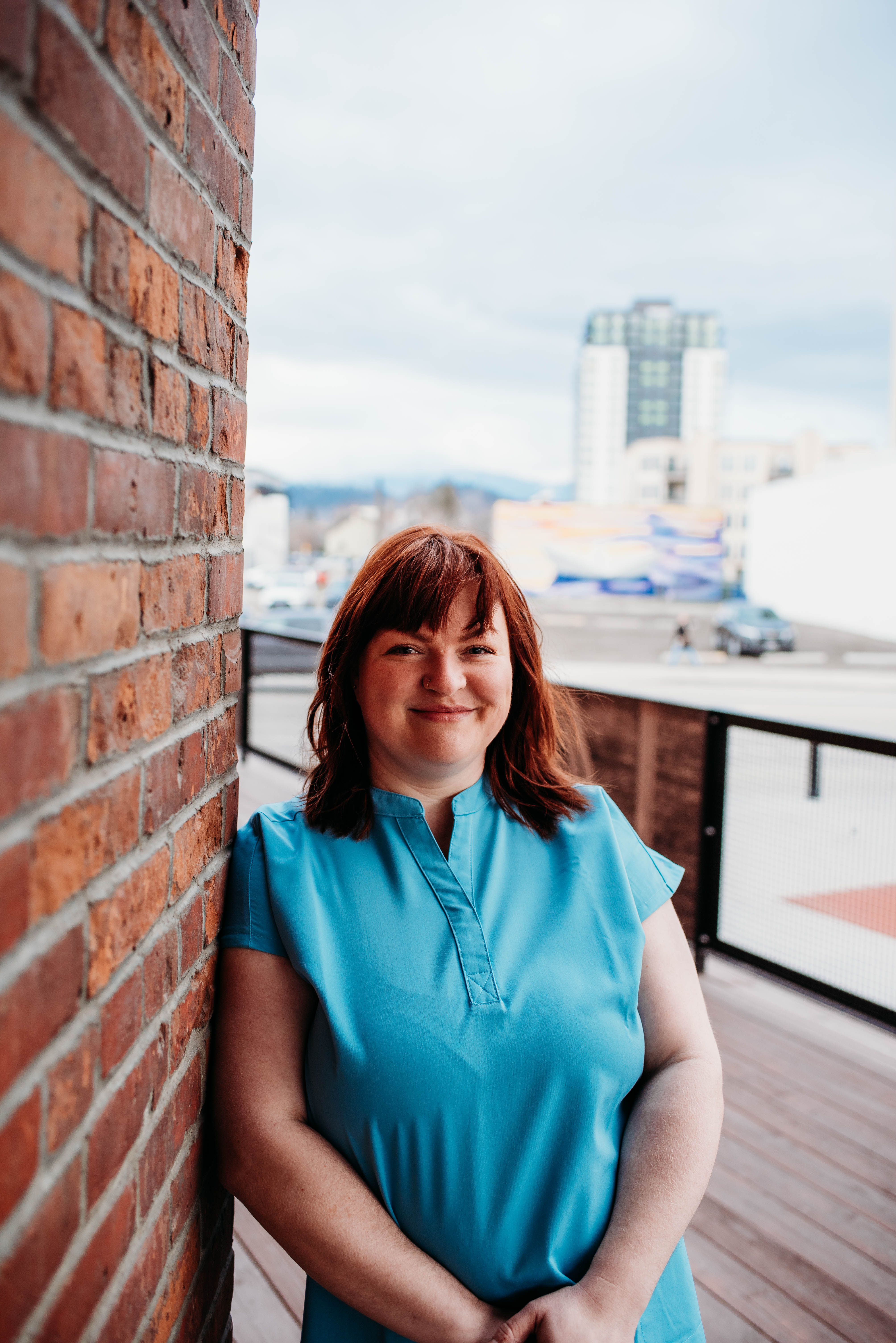 Smiling woman in a blue dress leaning against a brick wall with a cityscape background.