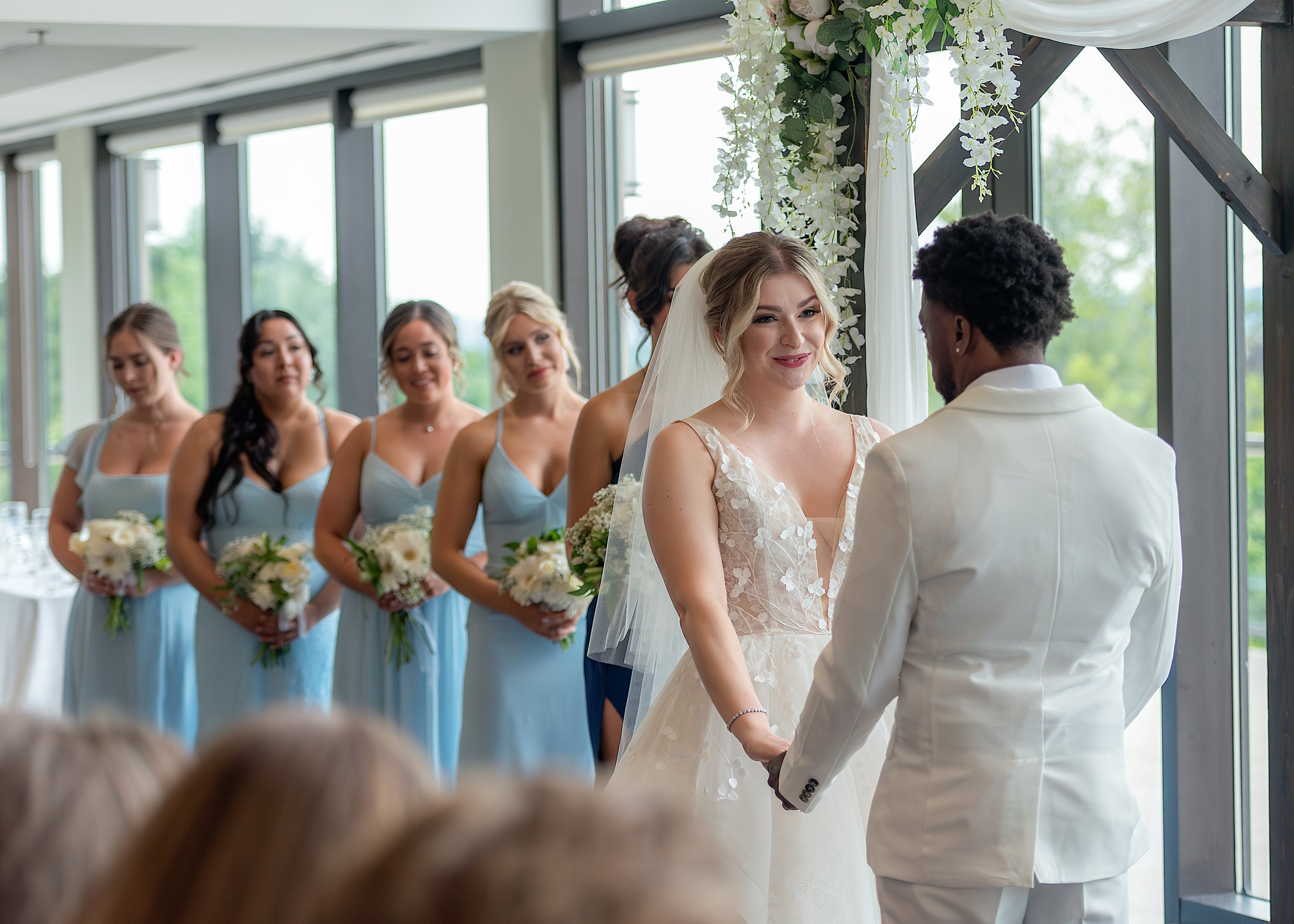 Bride and groom exchanging vows, surrounded by bridesmaids in blue dresses holding bouquets, under a floral arch in a bright venue.