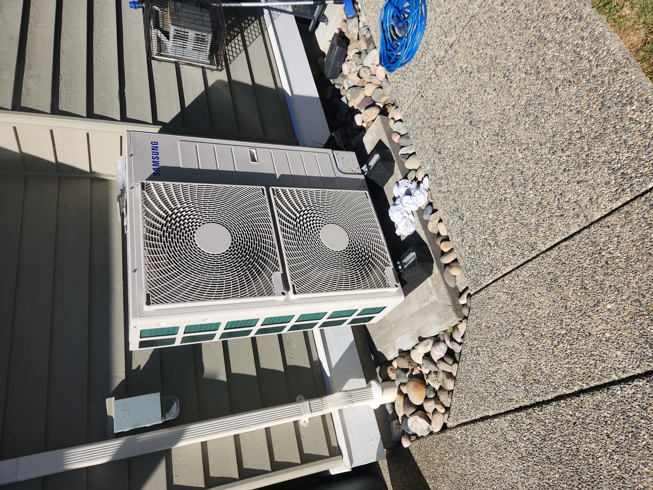 HVAC technician in blue uniform and yellow hard hat inspecting outdoor air conditioning unit using a tablet, with residential building and green garden in background.