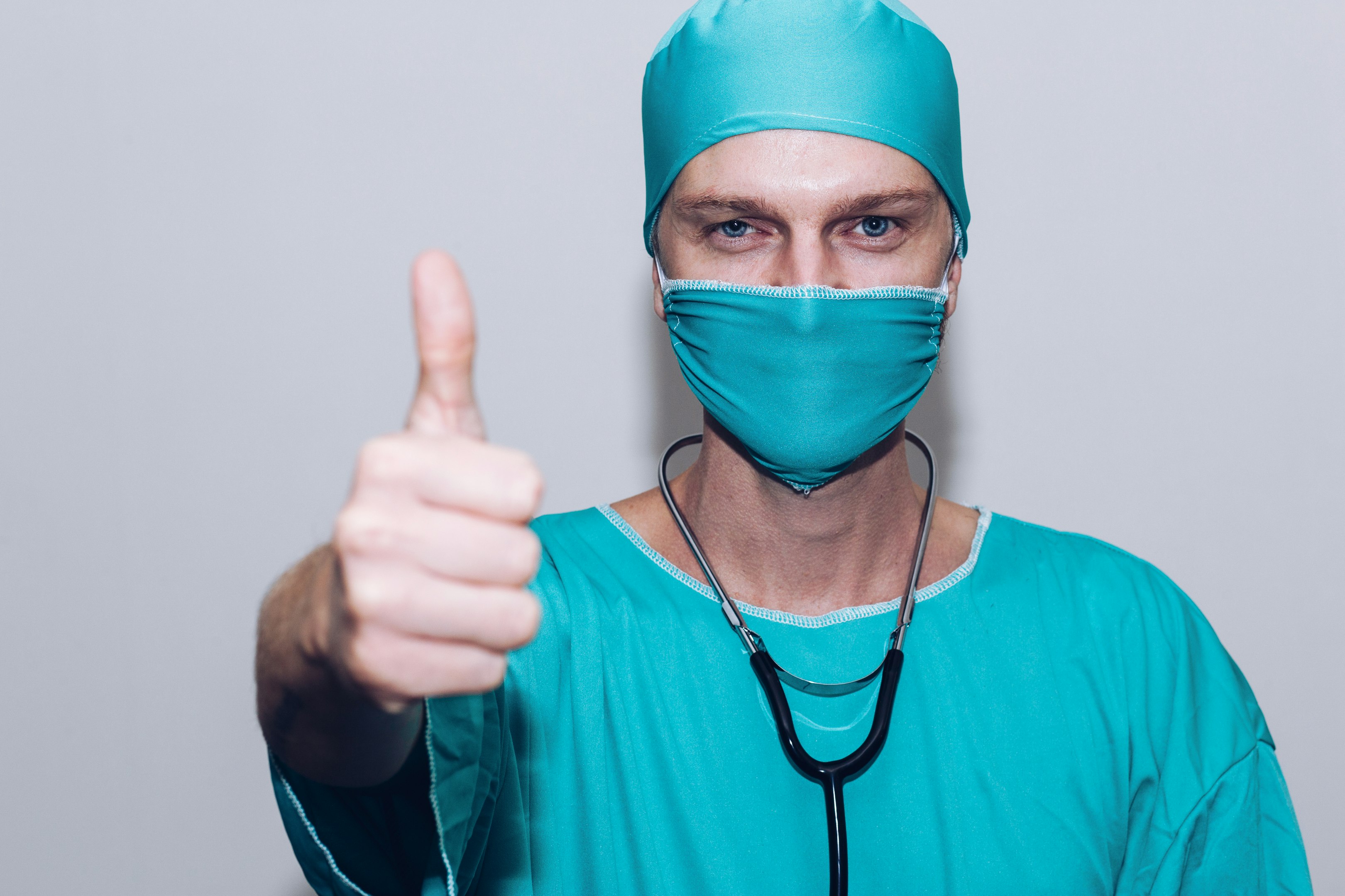 Surgeon wearing teal scrubs and mask giving a thumbs up with a stethoscope around neck.