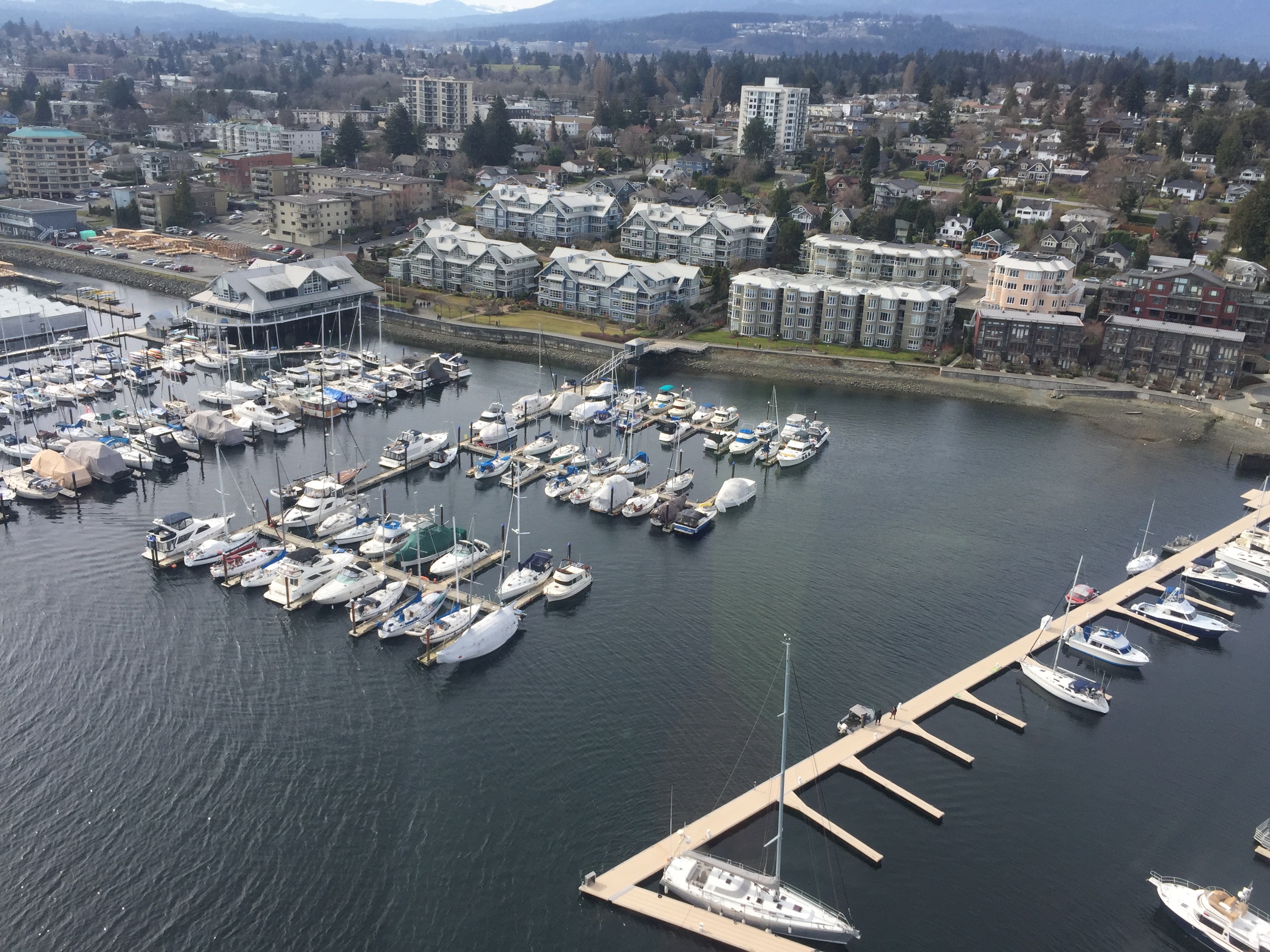 Aerial view of a marina with numerous yachts and boats docked, surrounded by residential buildings and a small harbor, with a backdrop of a scenic town and forested hills.