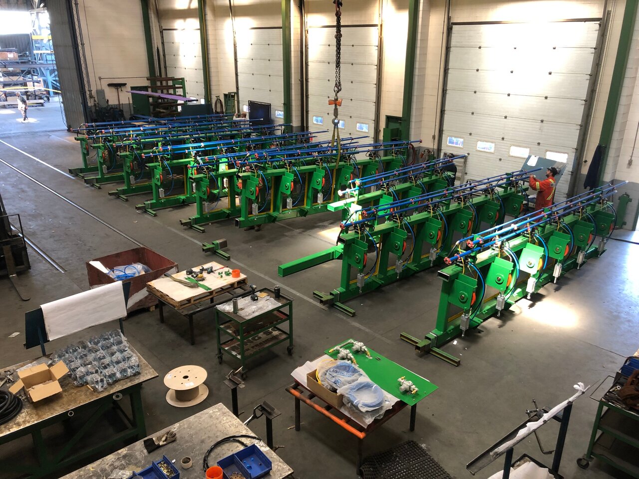 Industrial warehouse interior with multiple green machines lined up, workstations with tools and materials, and a worker inspecting equipment.